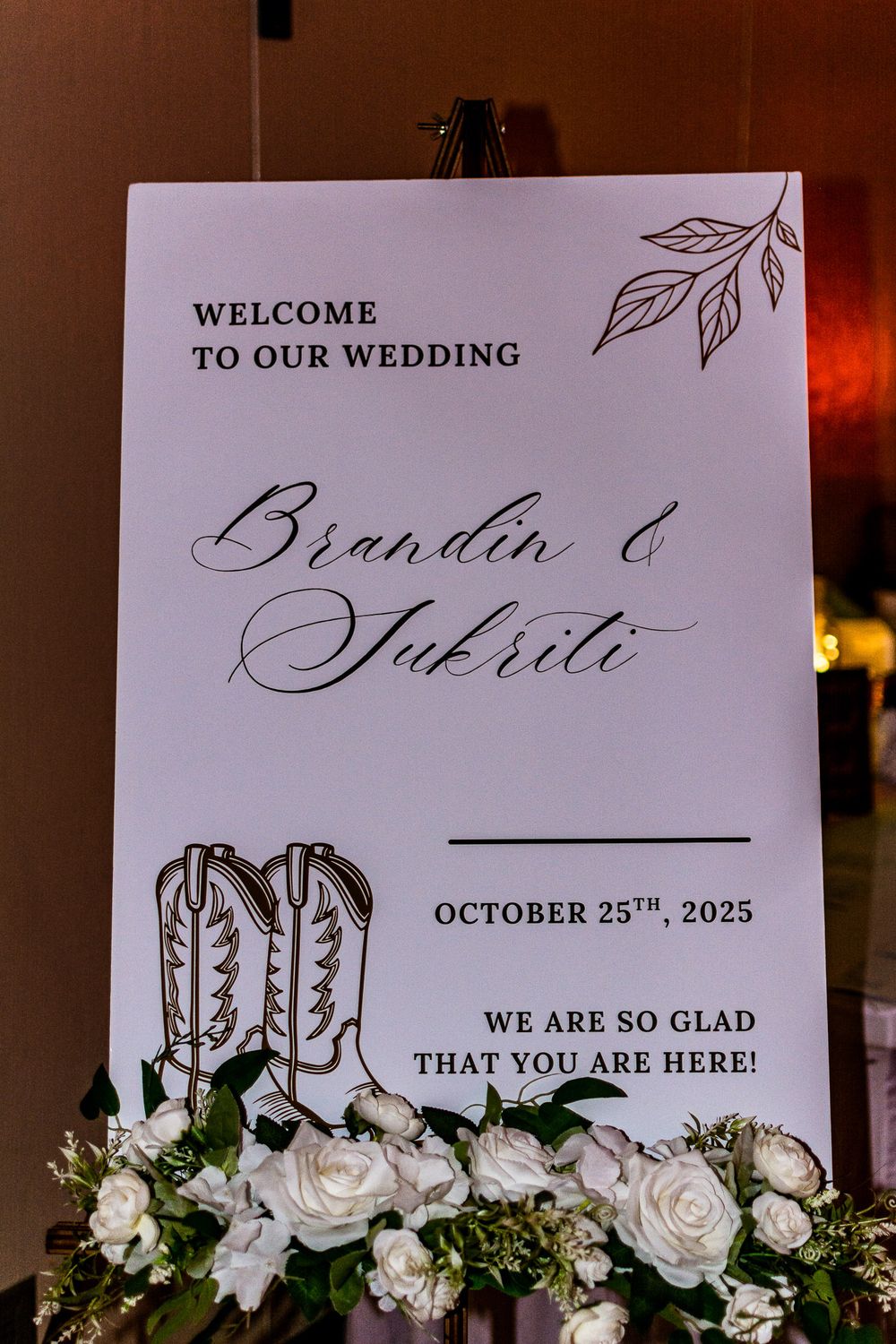 Wedding welcome sign with cowboy boot design and elegant calligraphy displayed among white roses and floral decorations.