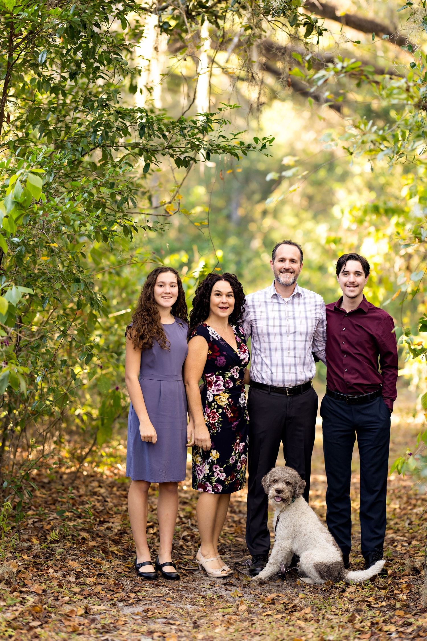 A group stands together on a leaf-covered path in an autumn setting with their fluffy white dog.