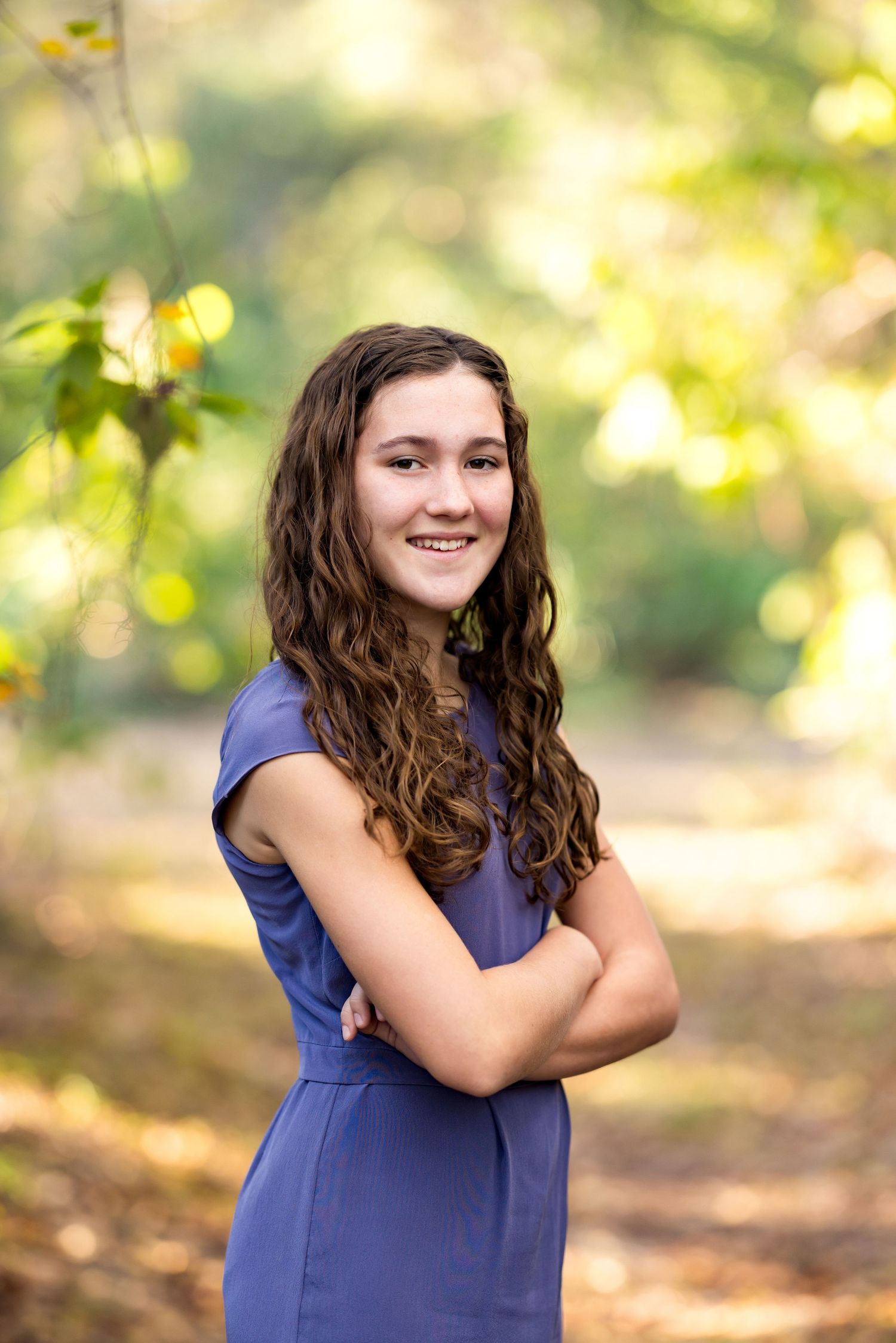 A young woman in a navy blue dress poses outdoors with crossed arms and long wavy hair against a blurred natural background.