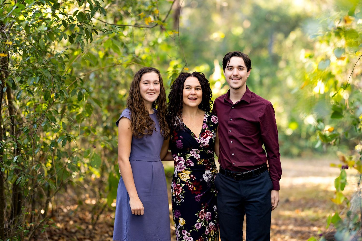 Three people stand together on a sunlit autumn path surrounded by green foliage.