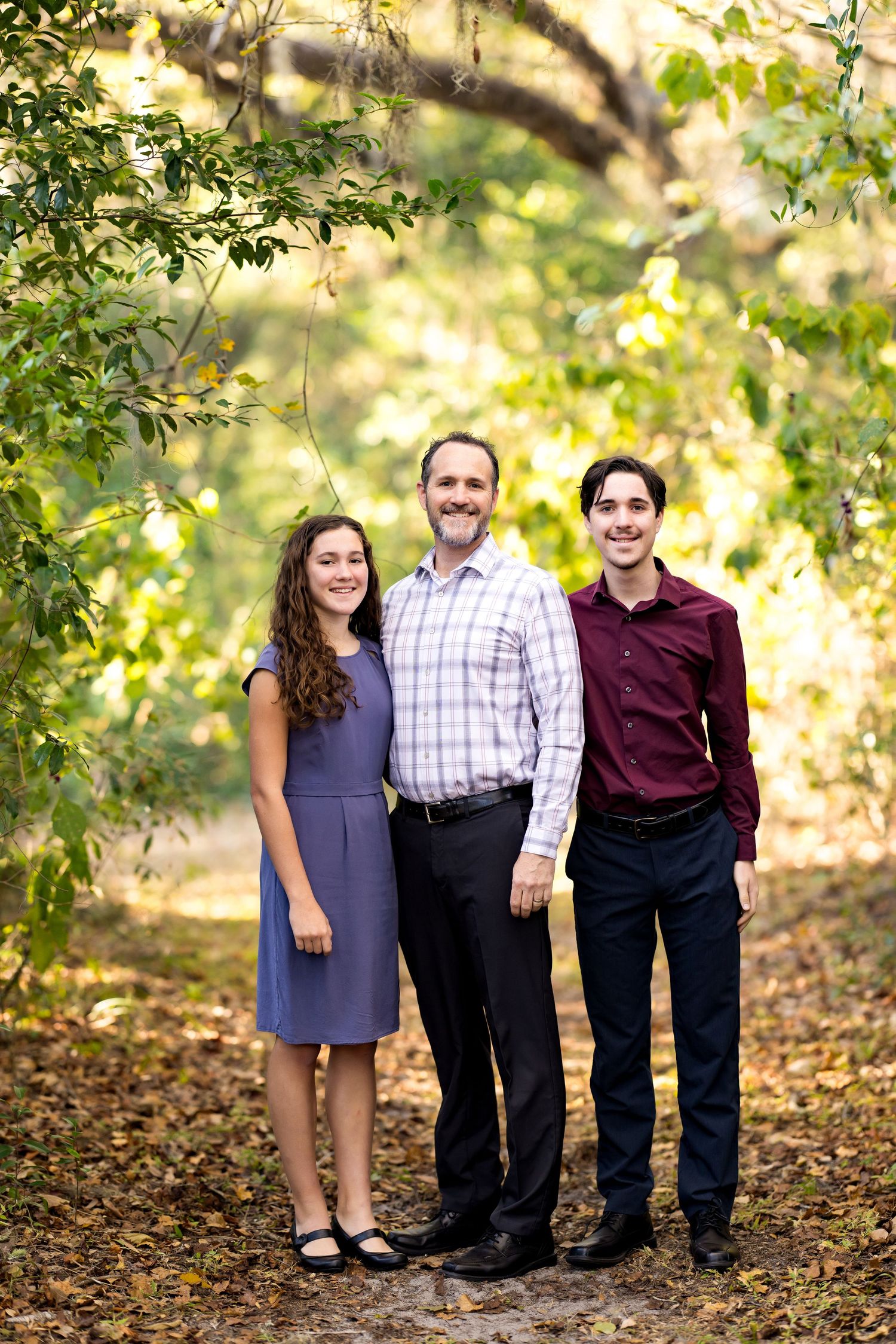 A family poses together in a wooded outdoor setting with golden sunlight filtering through the trees.
