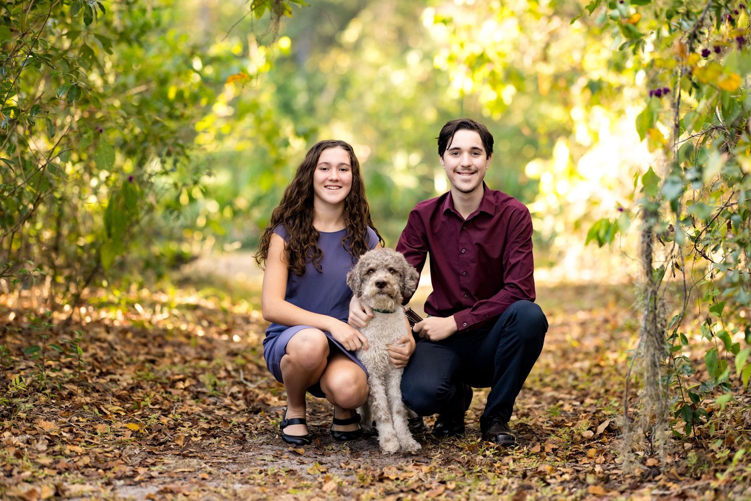 Two people kneeling on a leaf-covered path in autumn with a gray and white dog between them.