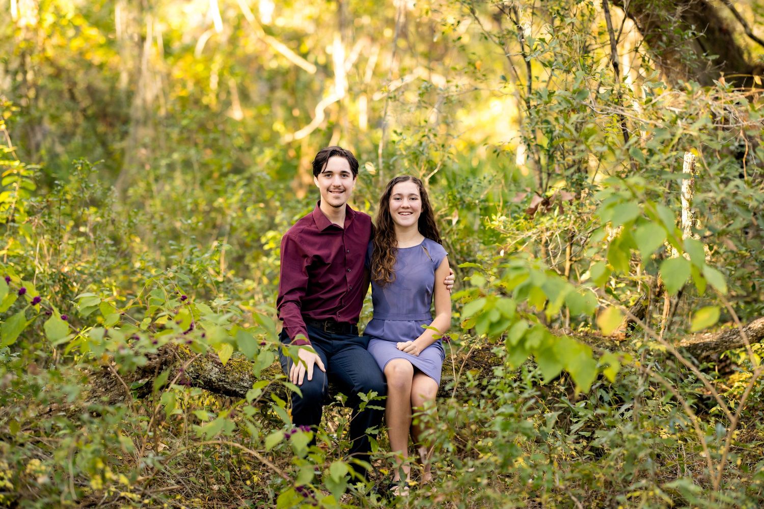 Two people sitting together outdoors in a sunlit wooded area with golden autumn foliage in the background.