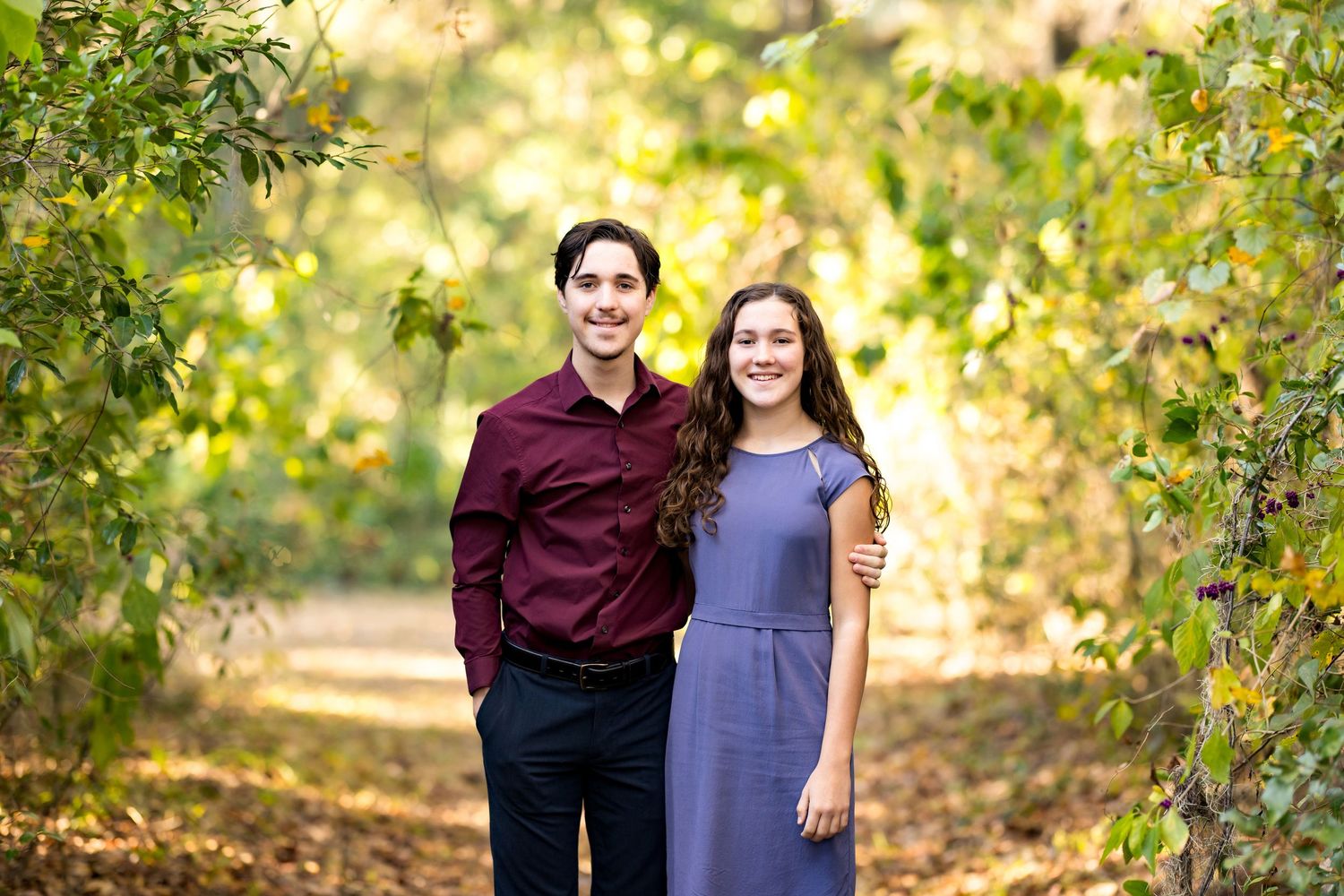 Two people stand together on a wooded path with golden sunlight filtering through autumn trees in the background.