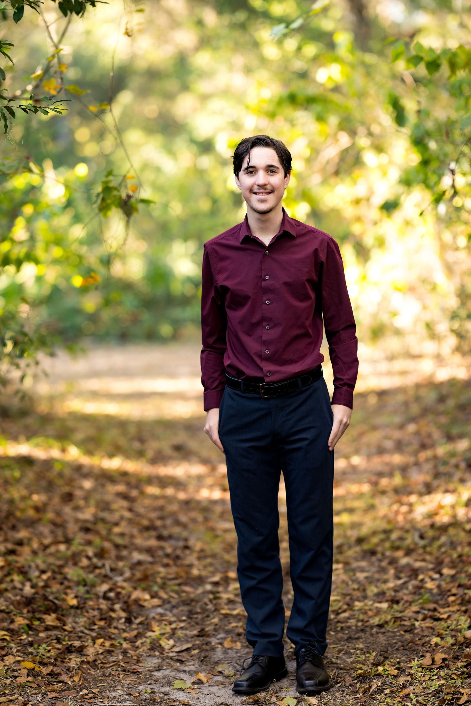 A person in a burgundy shirt and black pants stands on fallen autumn leaves in a sunlit wooded area.