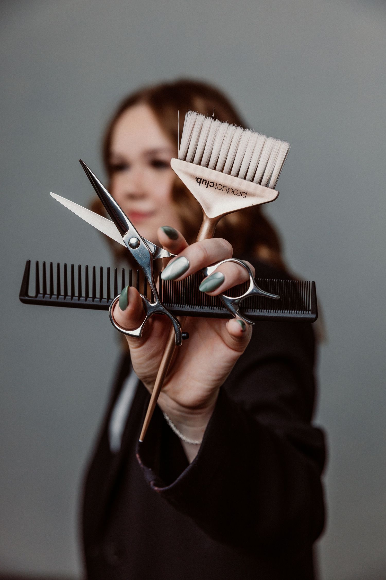 A female hairstylist holds scissors, combs, and a brush toward the camera in a professional studio setting.