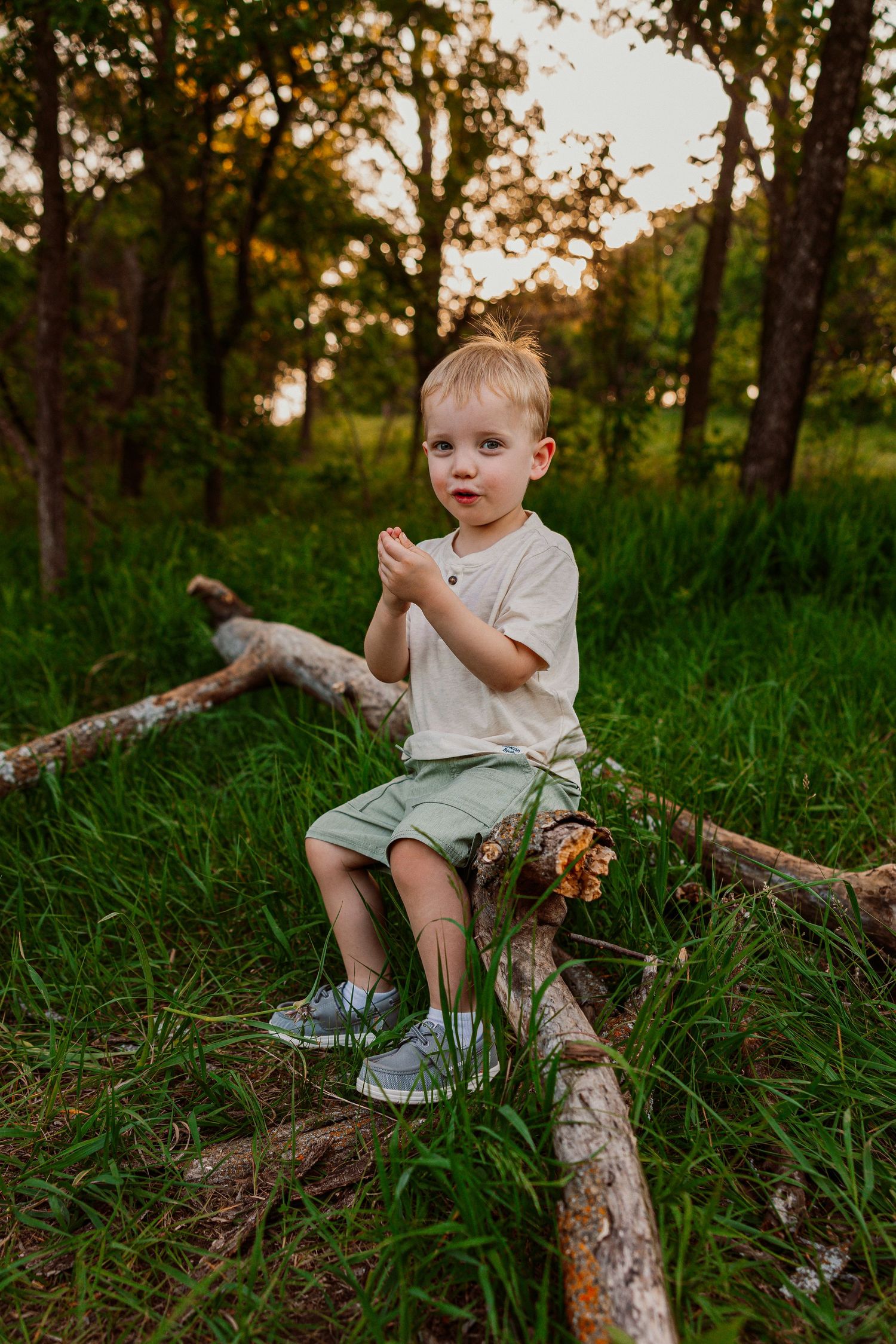 A young blonde boy sits on a fallen log in a lush green forest, clapping his hands at golden hour.