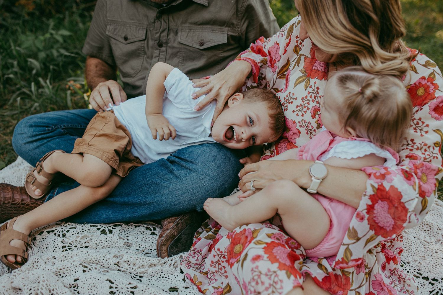 Family sitting on blanket outdoors, children laughing and playing in parents laps during a casual photo session.