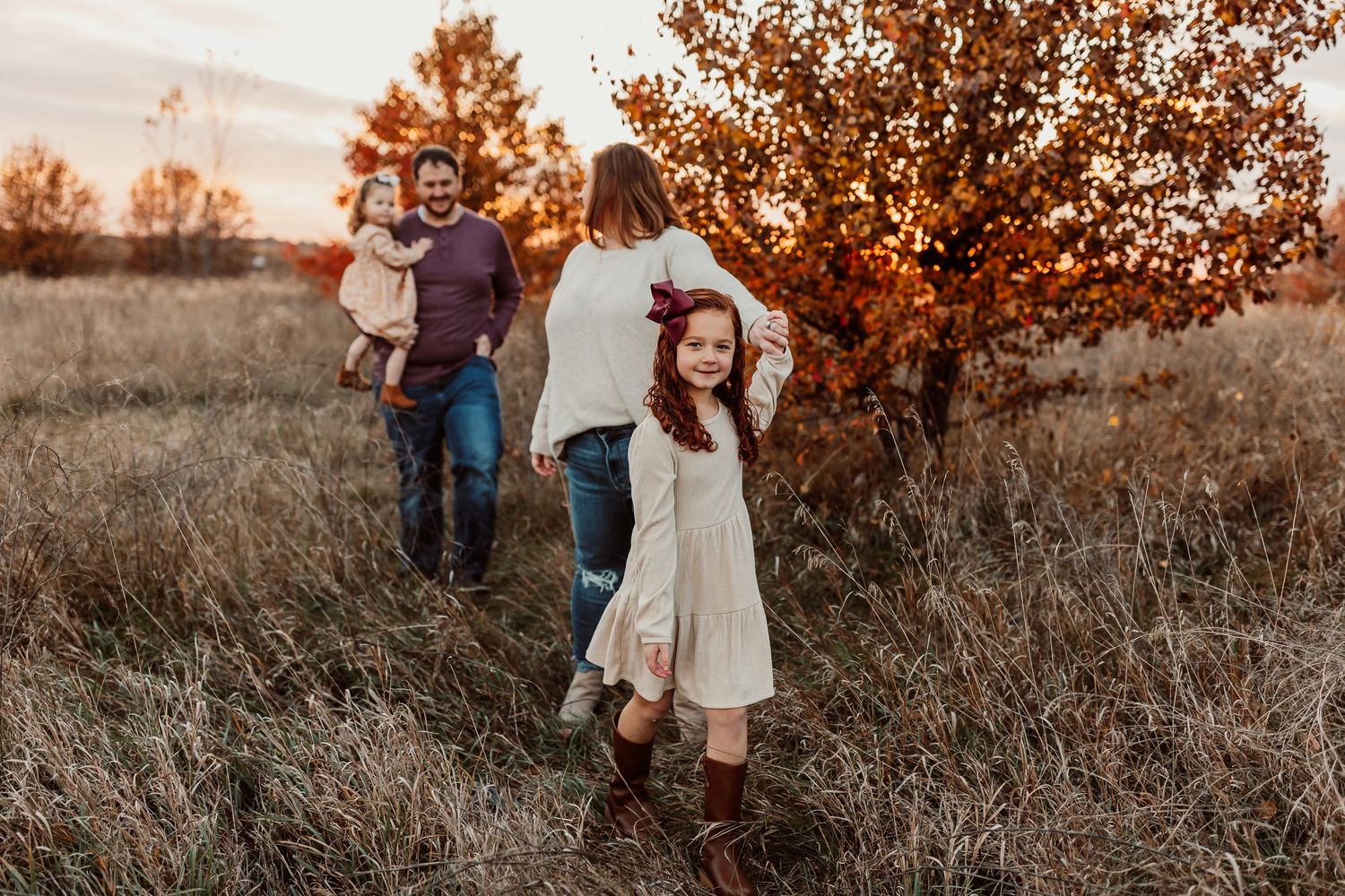 Family walking through autumn field at golden sunset, parents carrying child, young girl in white dress twirling near orange tree.