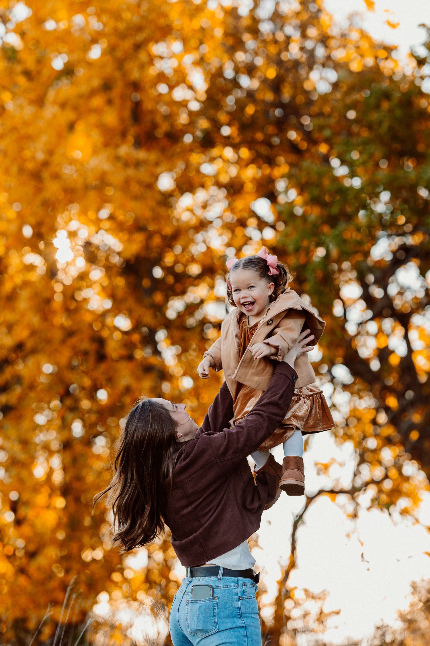 A mother joyfully lifts her laughing toddler against a vibrant autumn tree backdrop in golden fall colors.