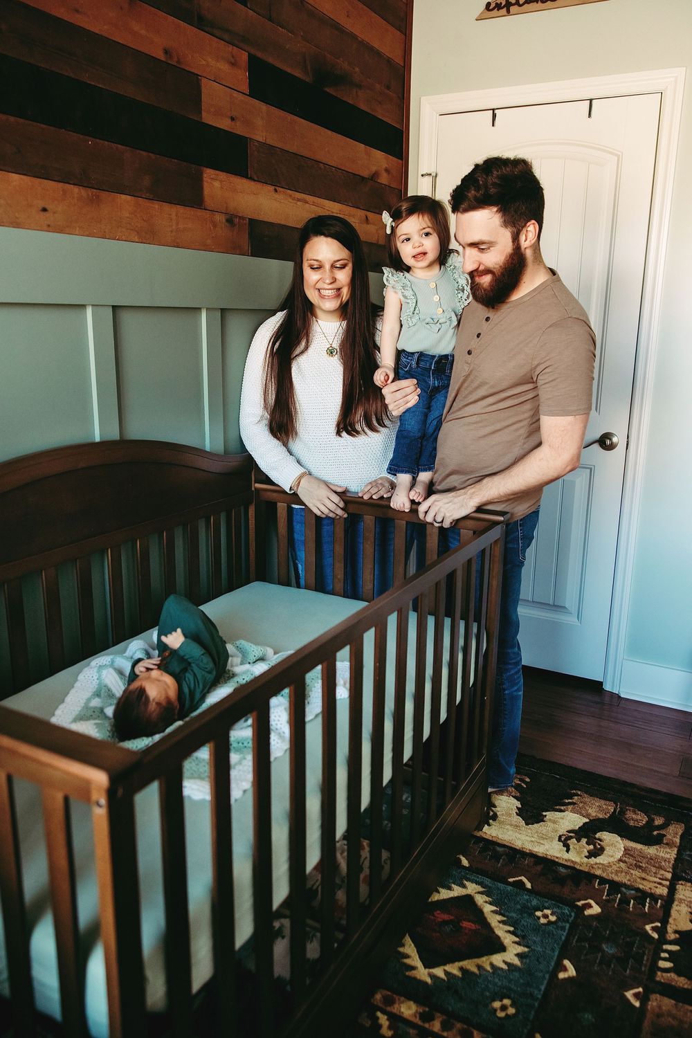 A family stands beside a wooden crib in a nursery with rustic wood paneled walls and patterned rug.