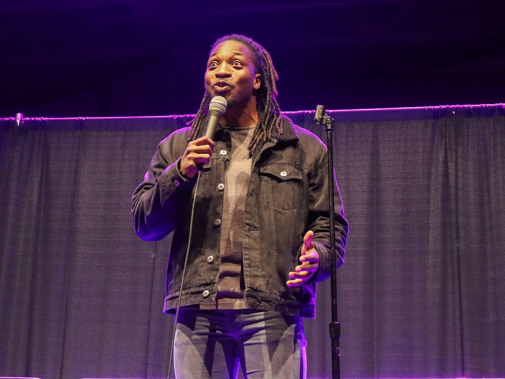 Stage performer in casual attire speaking into microphone under purple stage lighting.