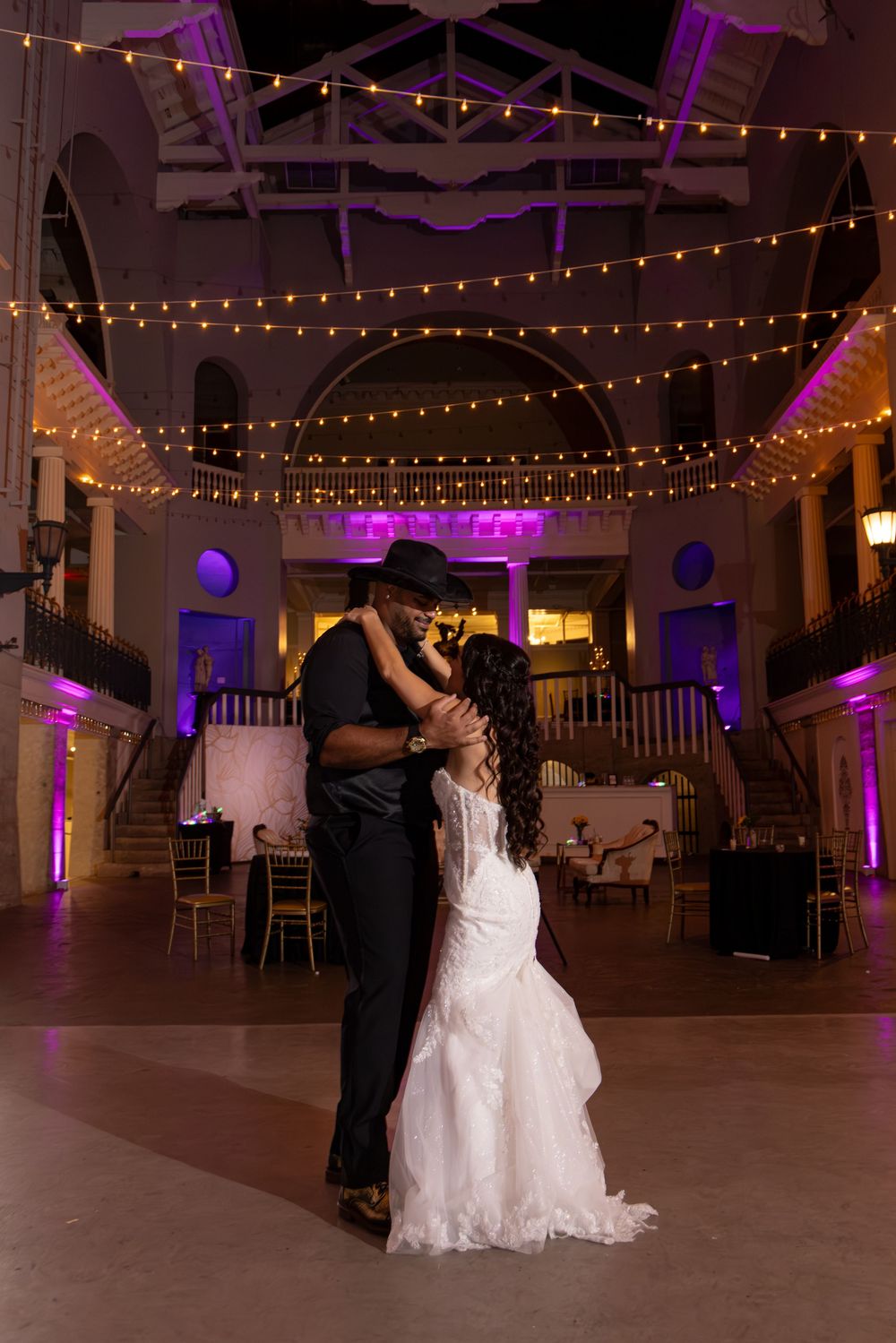 A couple dances together in an elegant reception hall decorated with string lights and purple uplighting.