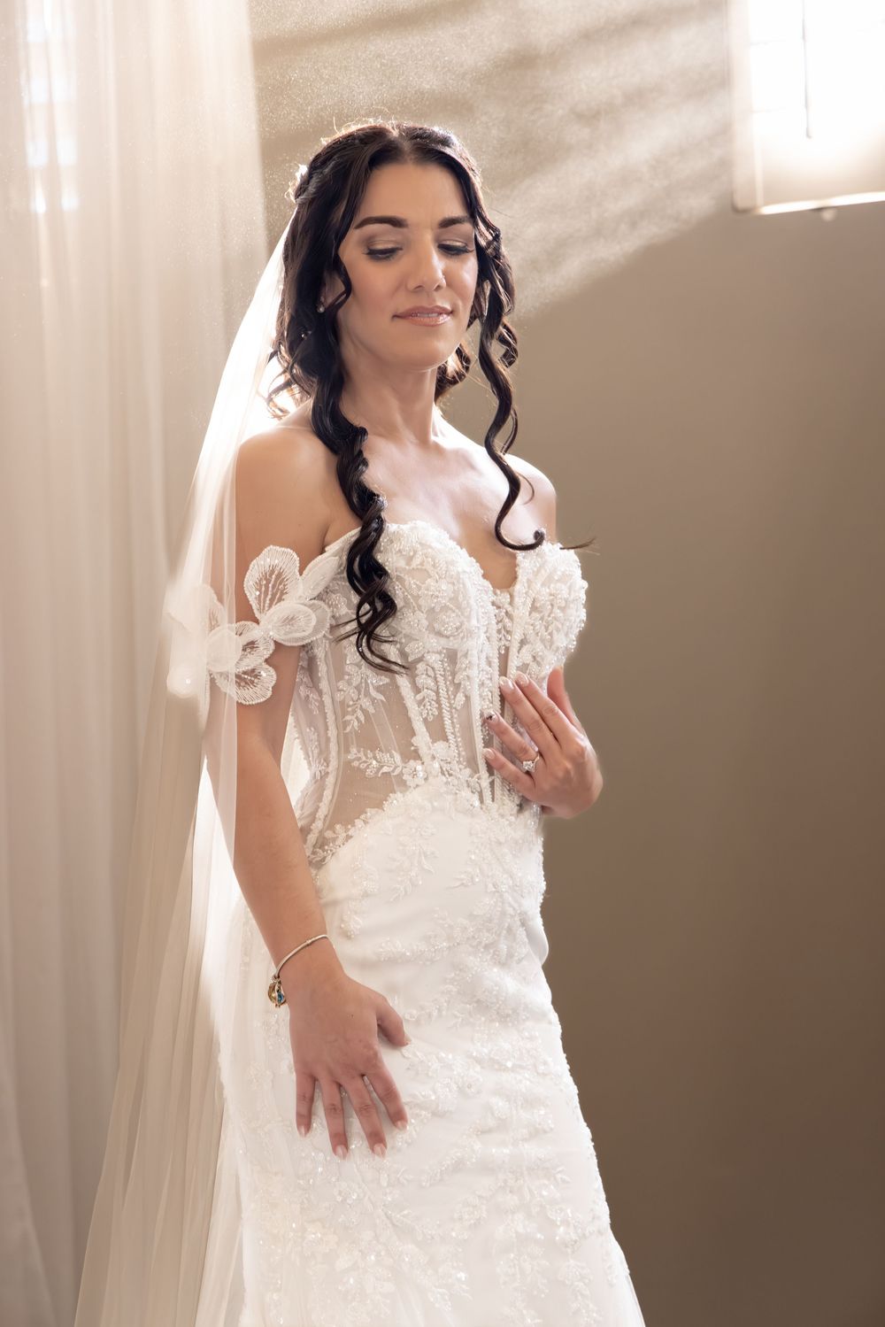 A bride in a white lace wedding dress poses gracefully in a softly lit room with romantic ambiance.