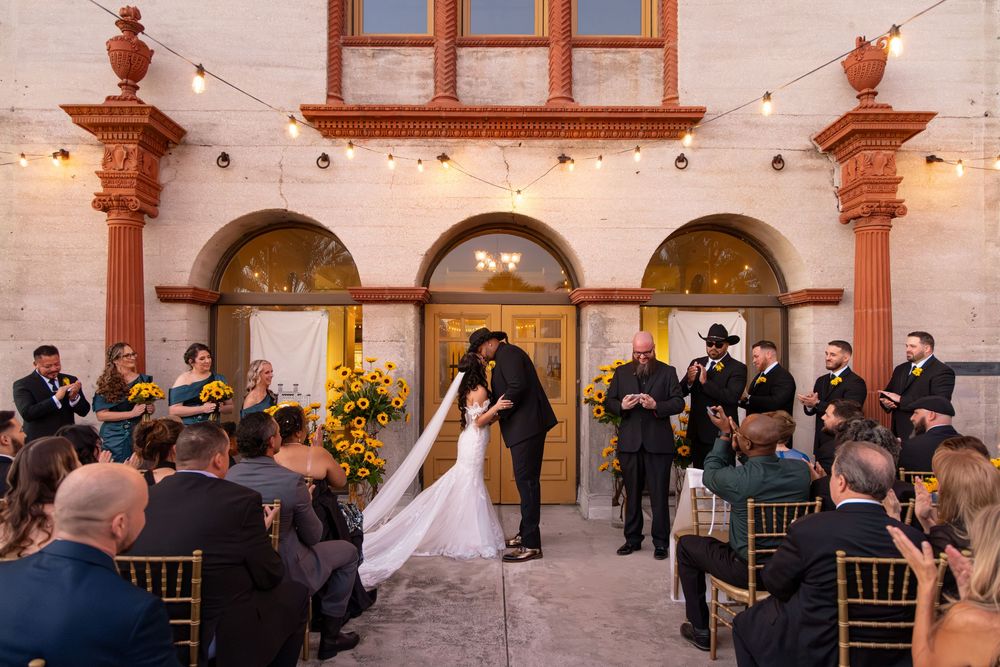 A wedding ceremony takes place under ornate archways at sunset with guests seated in gold chairs and musicians playing.