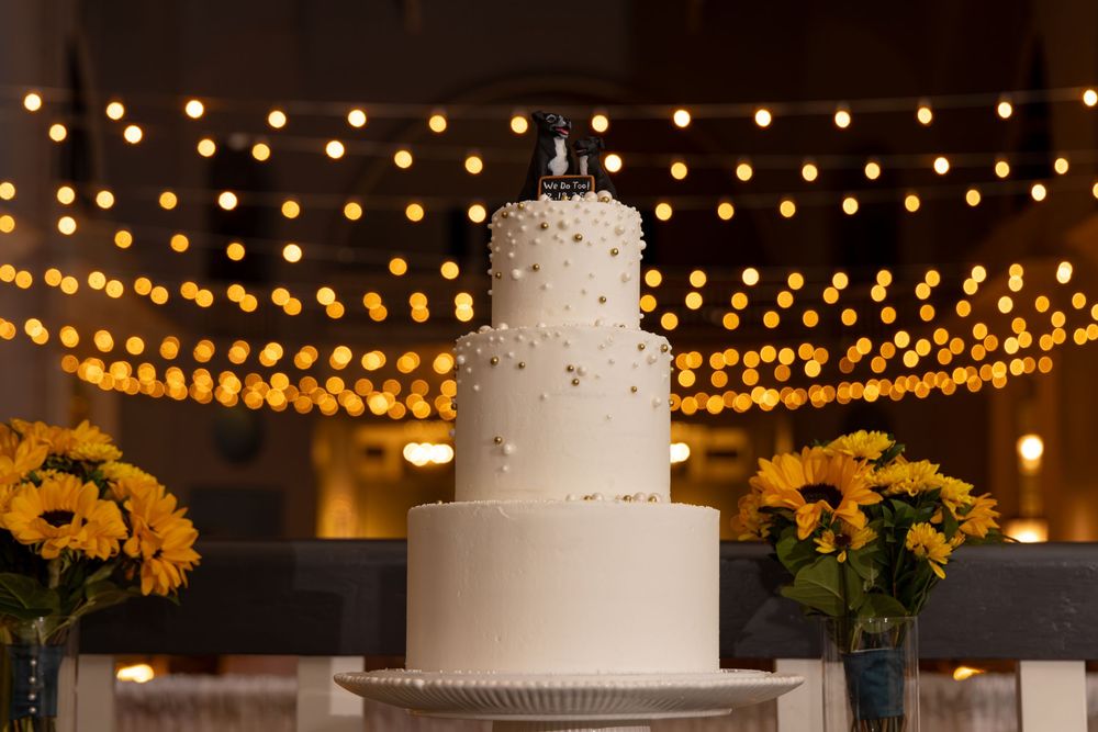 A white wedding cake decorated with delicate flowers sits beneath glowing string lights at an elegant reception venue.