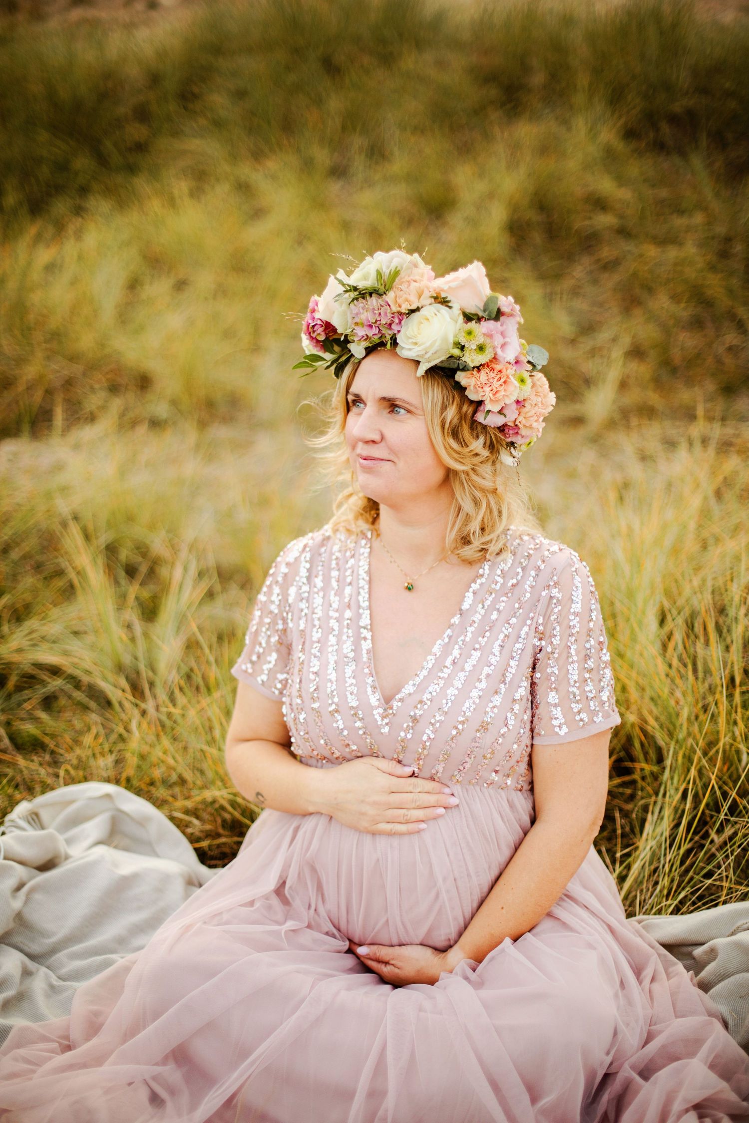 Blivande mamma i vacker klänning och blommig krona sitter fridfullt i högt gräs under gravidfotograferingen i vackra Ängelholm