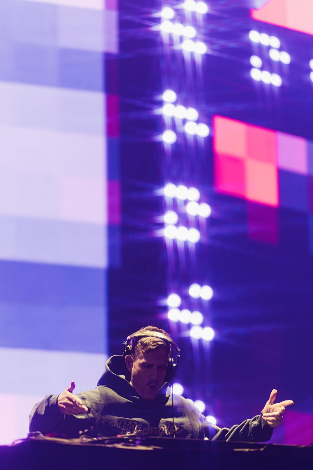 DJ performs behind decks at nightclub with colorful geometric lighting display glowing above in purple and pink tones.