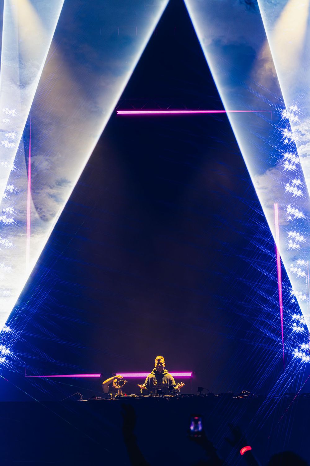 DJ performs on stage with dramatic triangular lighting display and blue spotlights creating an atmospheric concert scene.