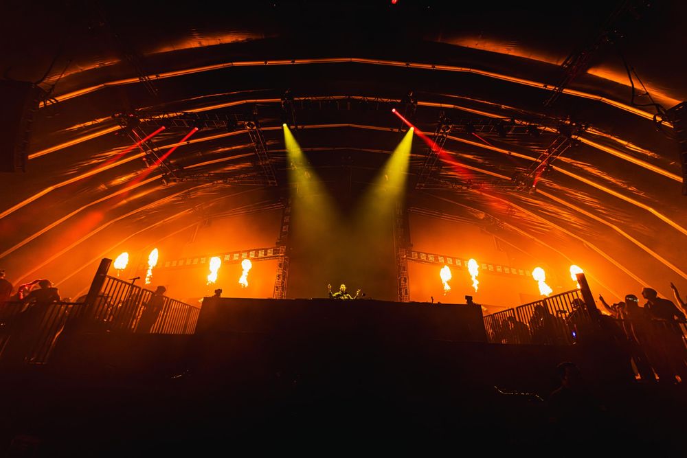 Concert stage illuminated with dramatic yellow spotlights and fire effects creating an orange glow across the arena.