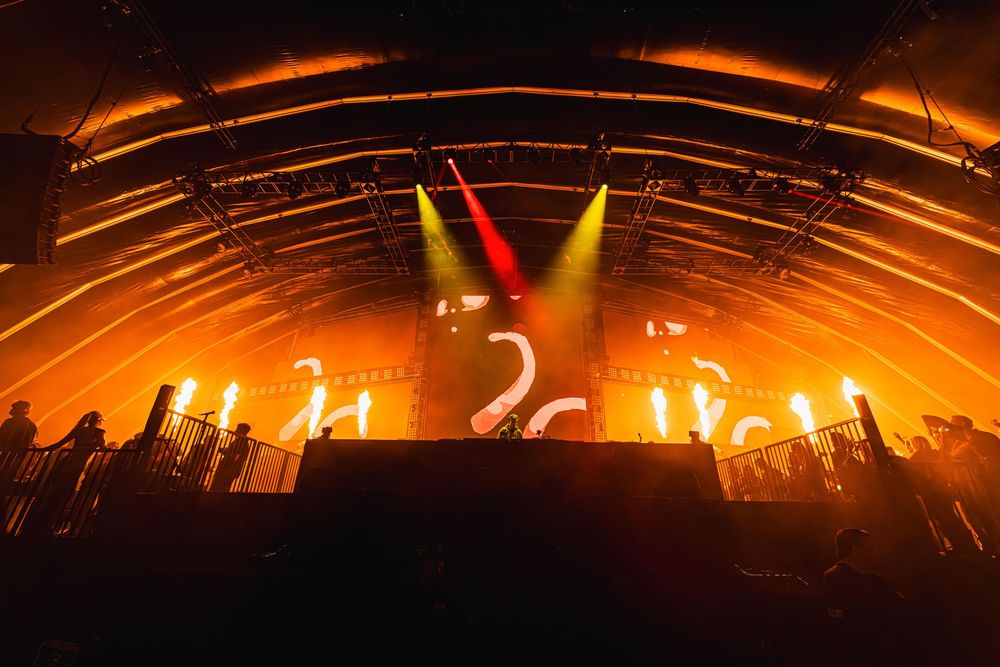 Dramatic orange stage lighting creates sweeping arcs across a dark concert venue ceiling during a live performance.