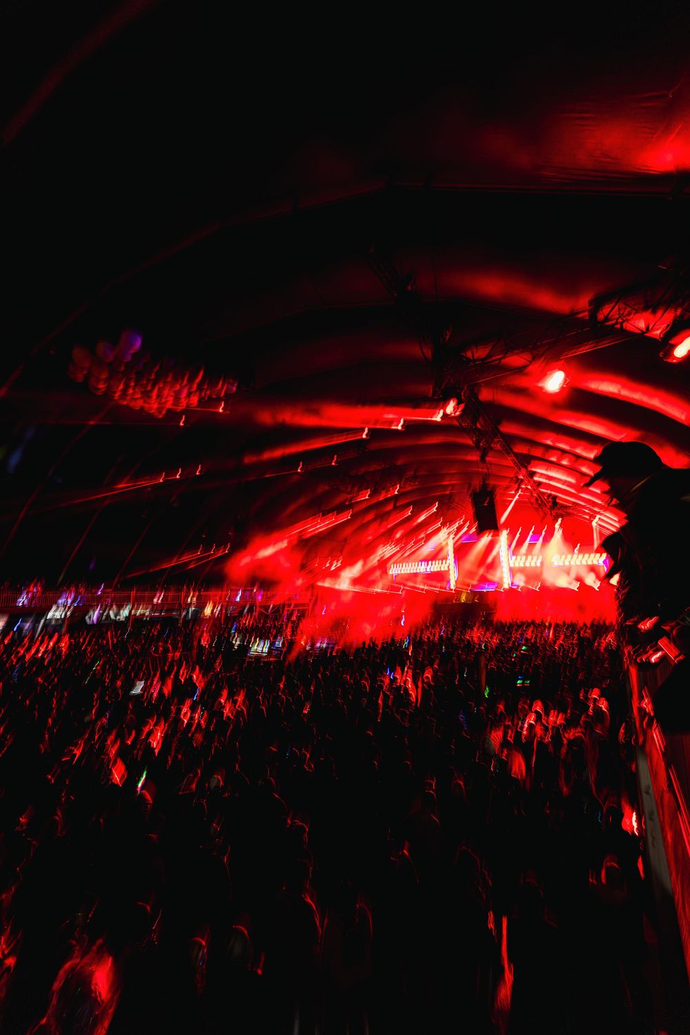 Large concert crowd illuminated by dramatic red lighting and stage effects at a nighttime music performance venue.