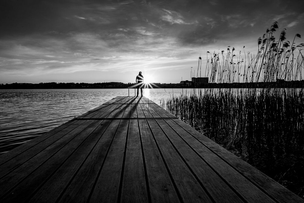 Sort/hvidt fotografi af en person der står i silhuet for enden af en badebro og kigger på solnedgangen og Viborg.