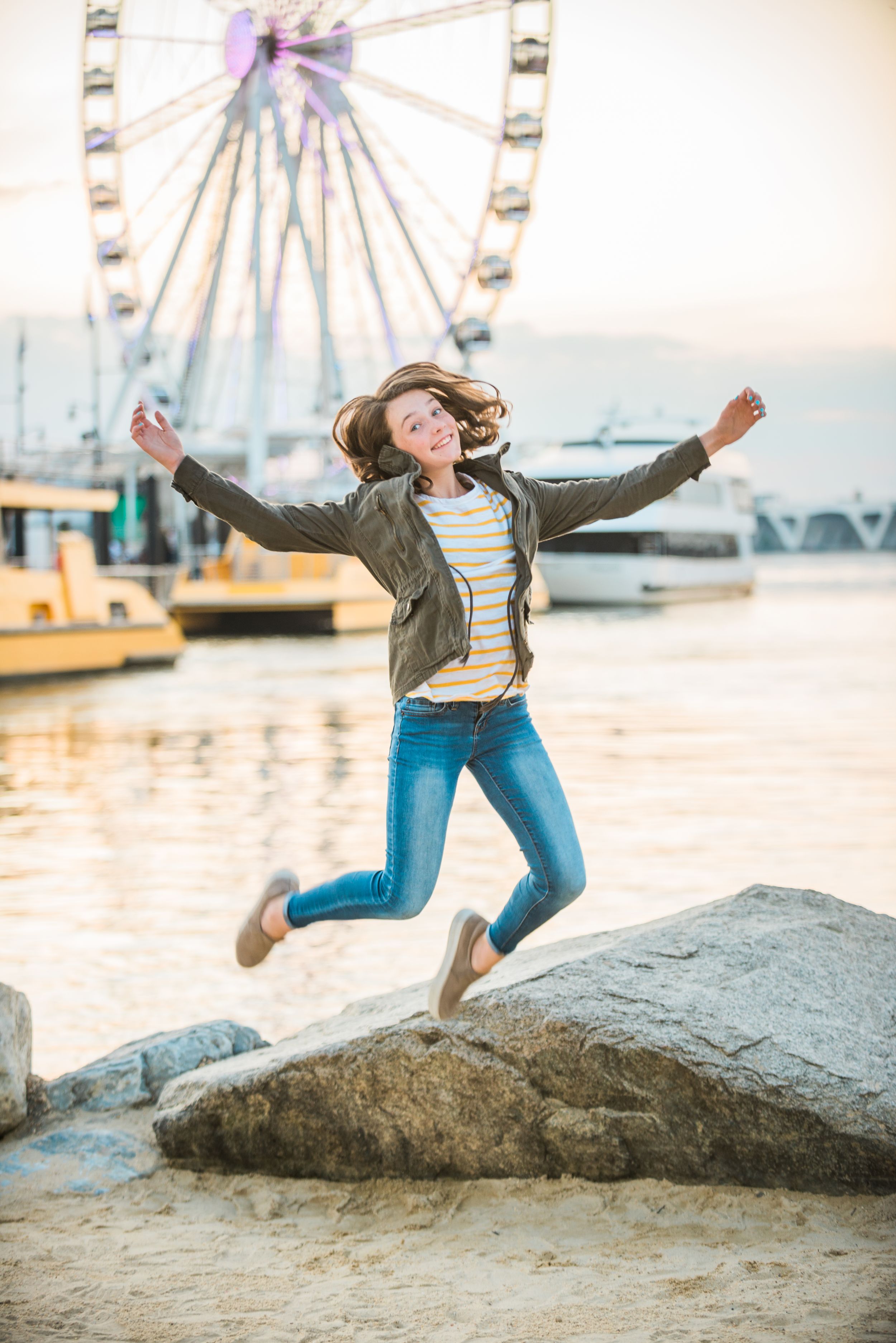 Person jumping joyfully on rocks near a ferris wheel at sunset.