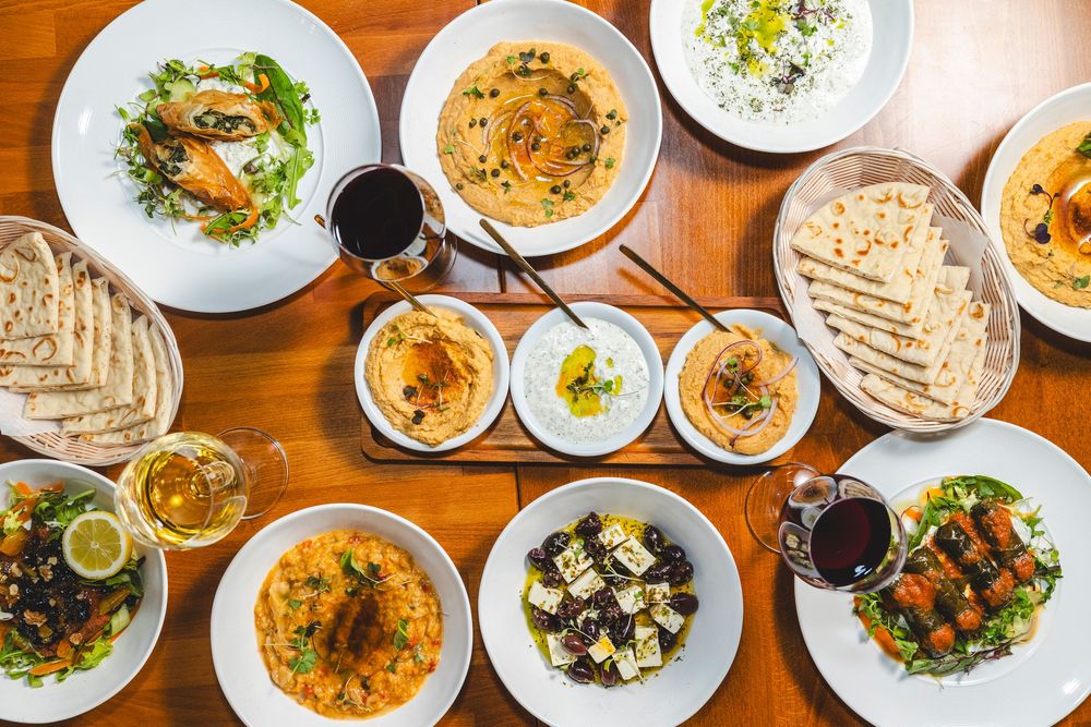 An overhead view of a wooden table set with multiple plates of Mediterranean dishes and wine glasses.