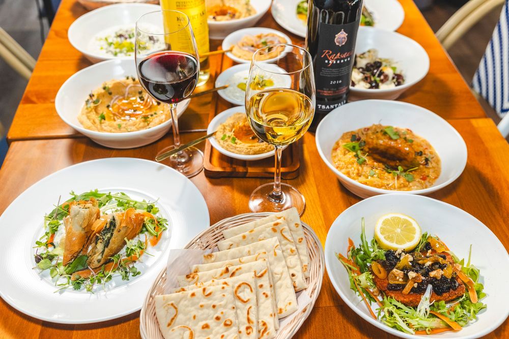 Mediterranean feast spread on wooden table with wine glasses, flatbread, salads and various entrees.