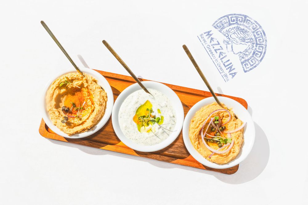 Three creamy dips served in white bowls on a wooden serving board with a blue restaurant stamp visible.