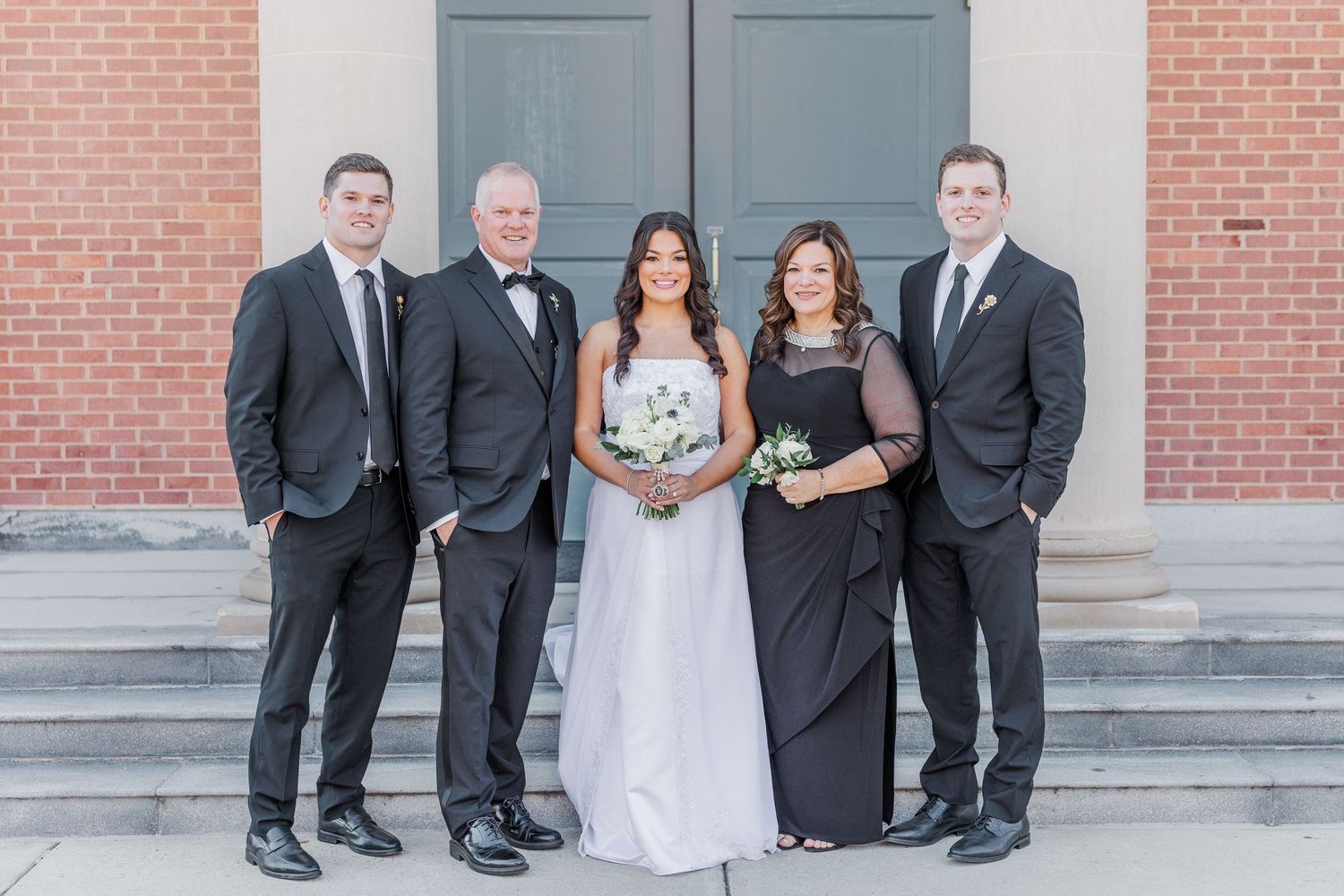 Wedding party in formal attire poses in front of brick building with classic architecture details.