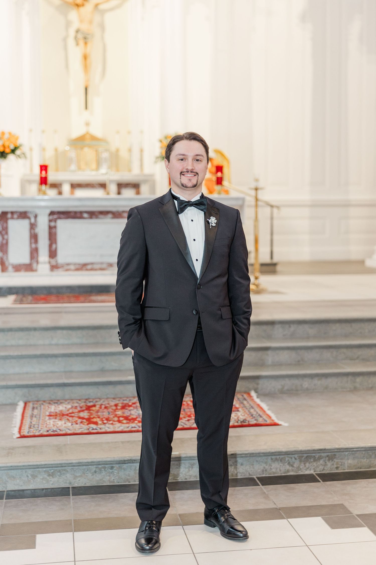 A groom in a classic black tuxedo stands inside an elegant white church with red accents and tiered steps.