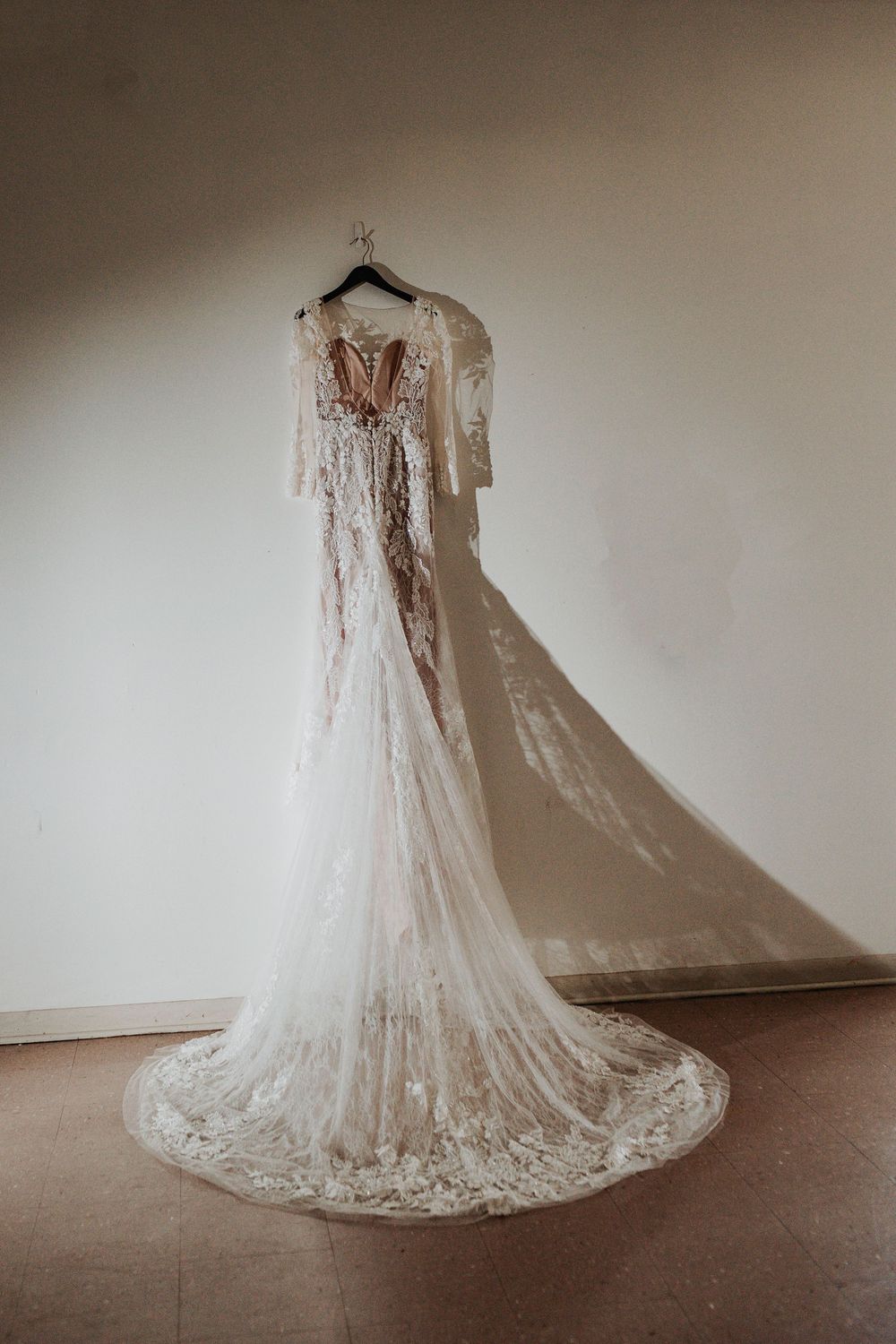 Elegant lace wedding dress with long sleeves and train hanging on display against neutral wall.
