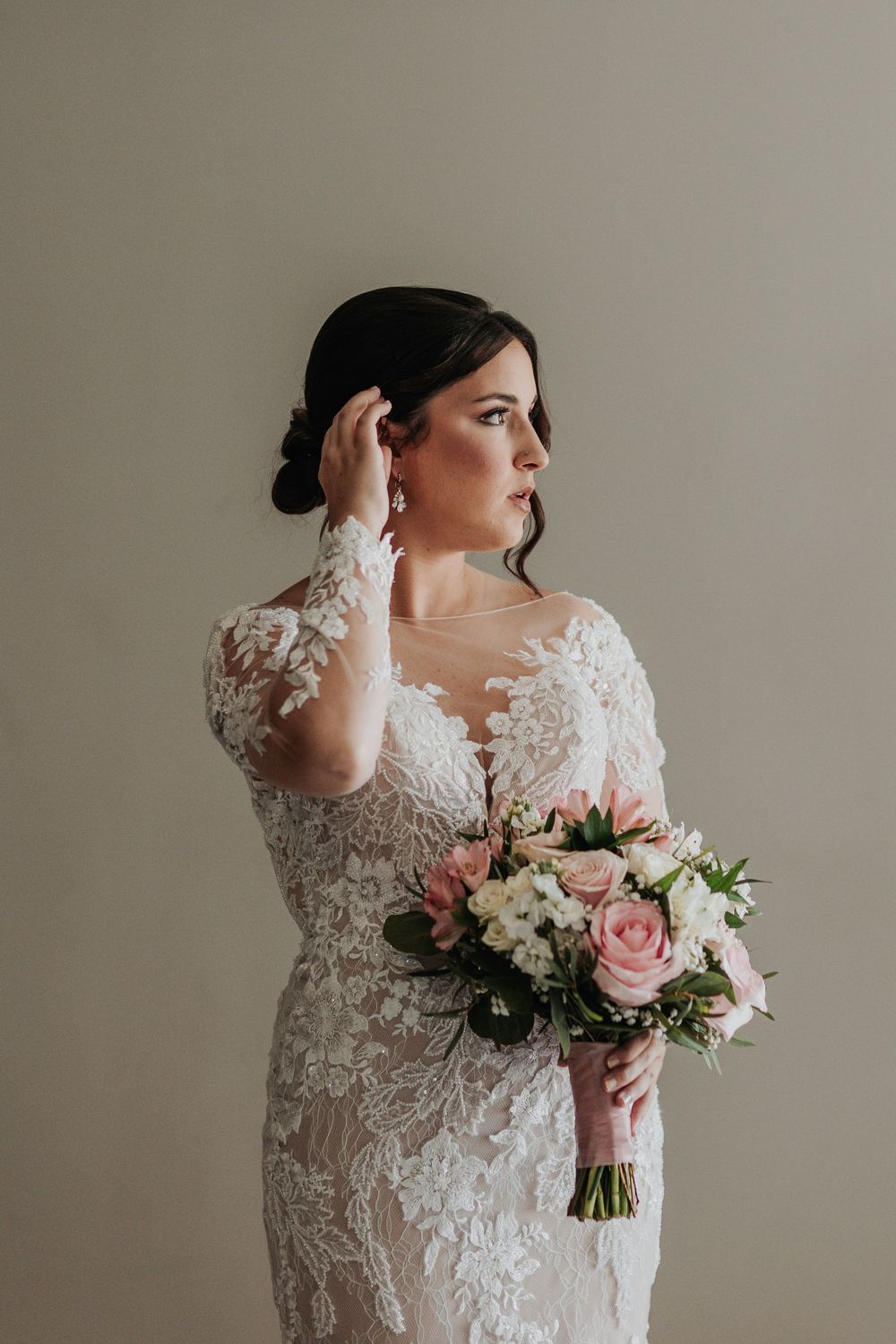 Bride in lace gown holds pink and white rose bouquet while touching her hair.