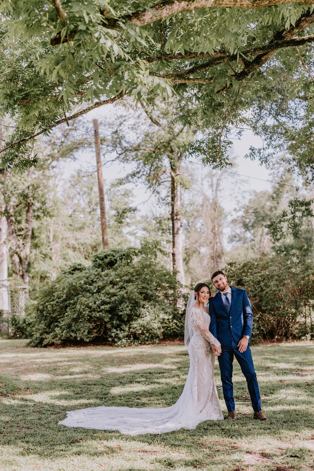 Wedding portrait in lush garden setting with tall trees and flowing bridal train.