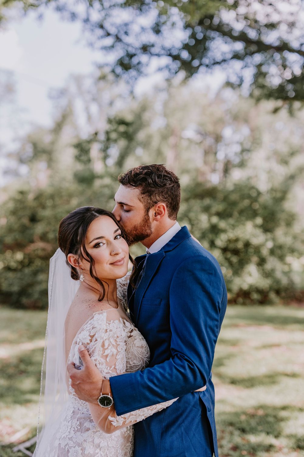 Tender moment between couple in sunny garden wearing navy suit and lace wedding gown.