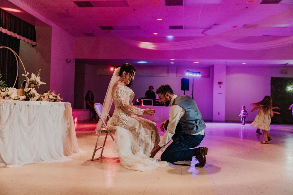 Wedding reception scene with pink lighting as couple participates in shoe game tradition.