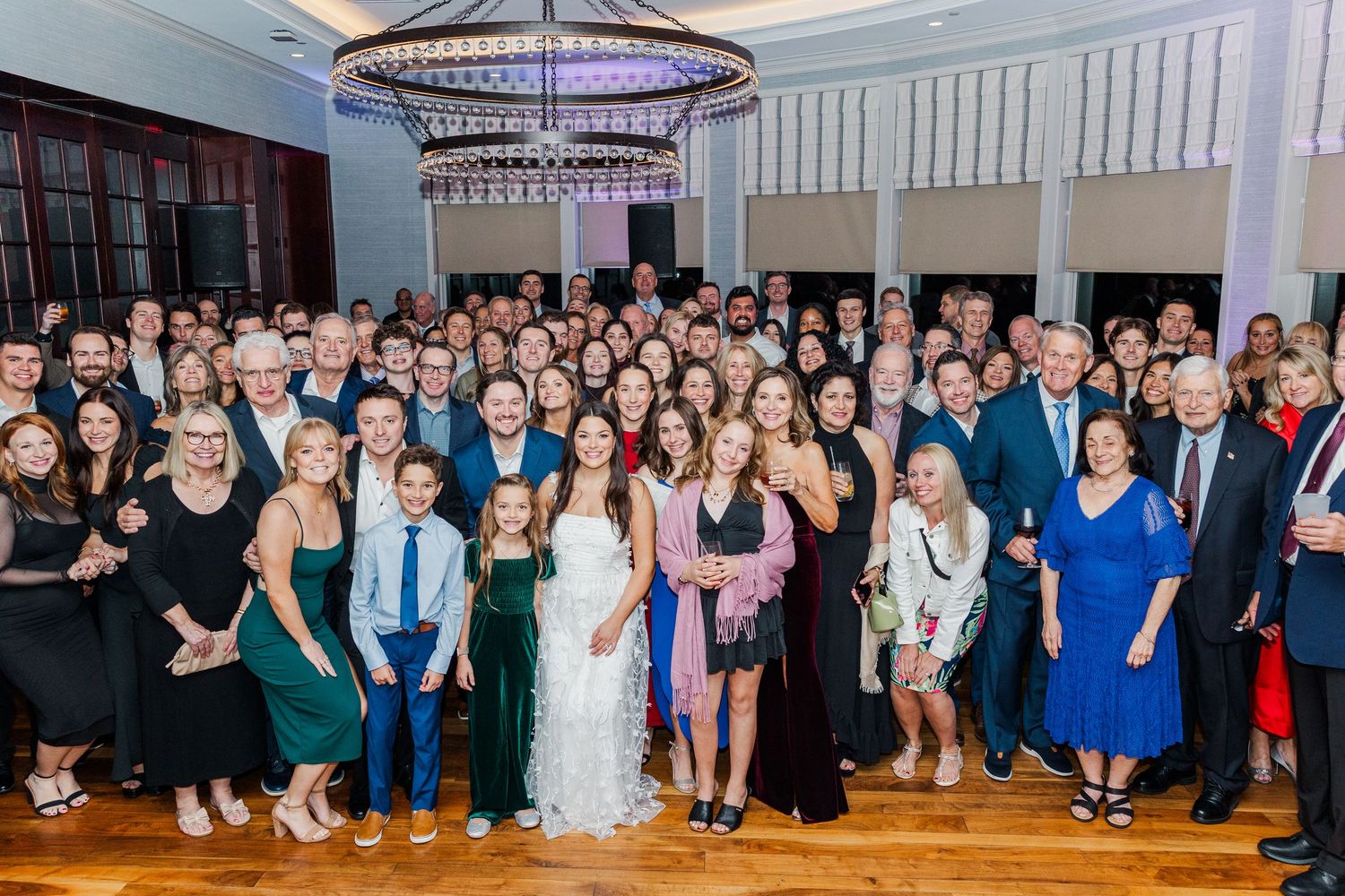 Large panoramic photo of wedding guests gathered in a reception hall under circular chandeliers.