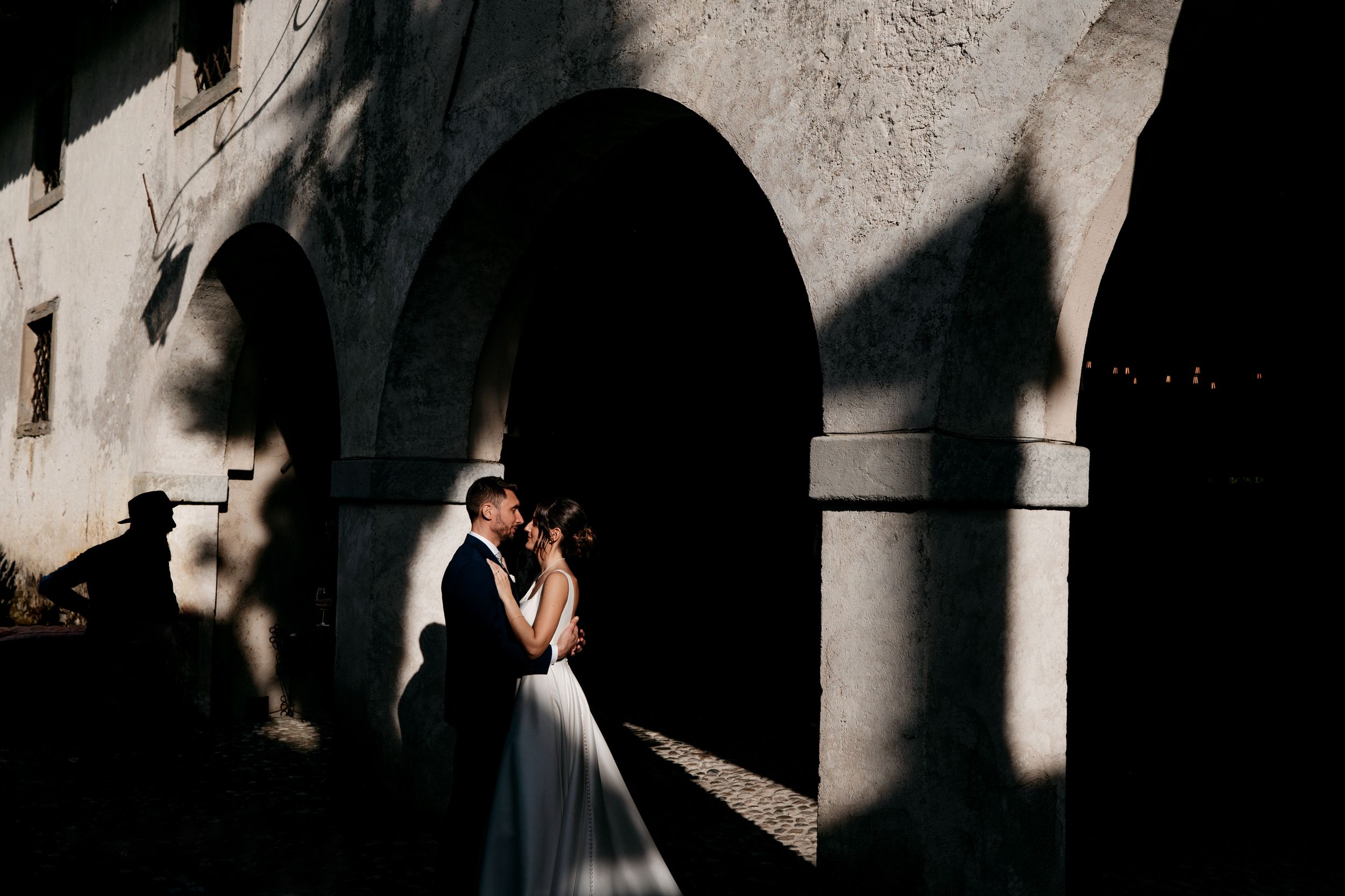 Elegant wedding couple embraces in dramatic lighting under stone archways during romantic photo session.