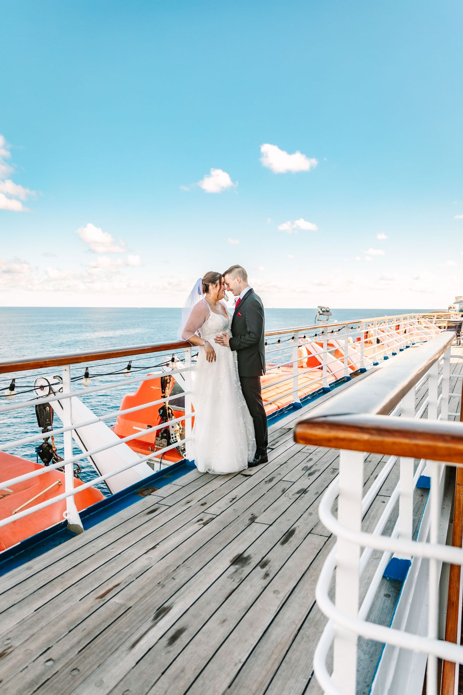 A couple in wedding attire shares an intimate moment on a cruise ship deck with bright orange benches and blue sky.