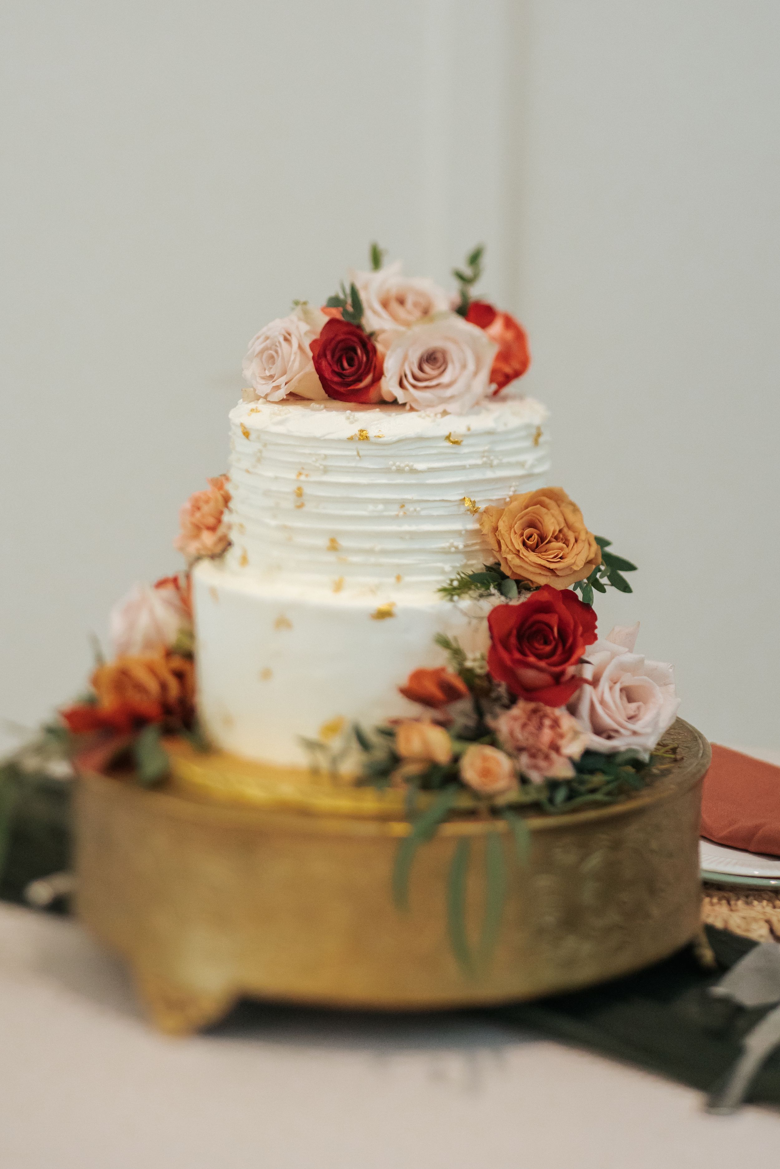 Elegant white wedding cake decorated with peach and red roses on a gold stand captured in multiple angles.