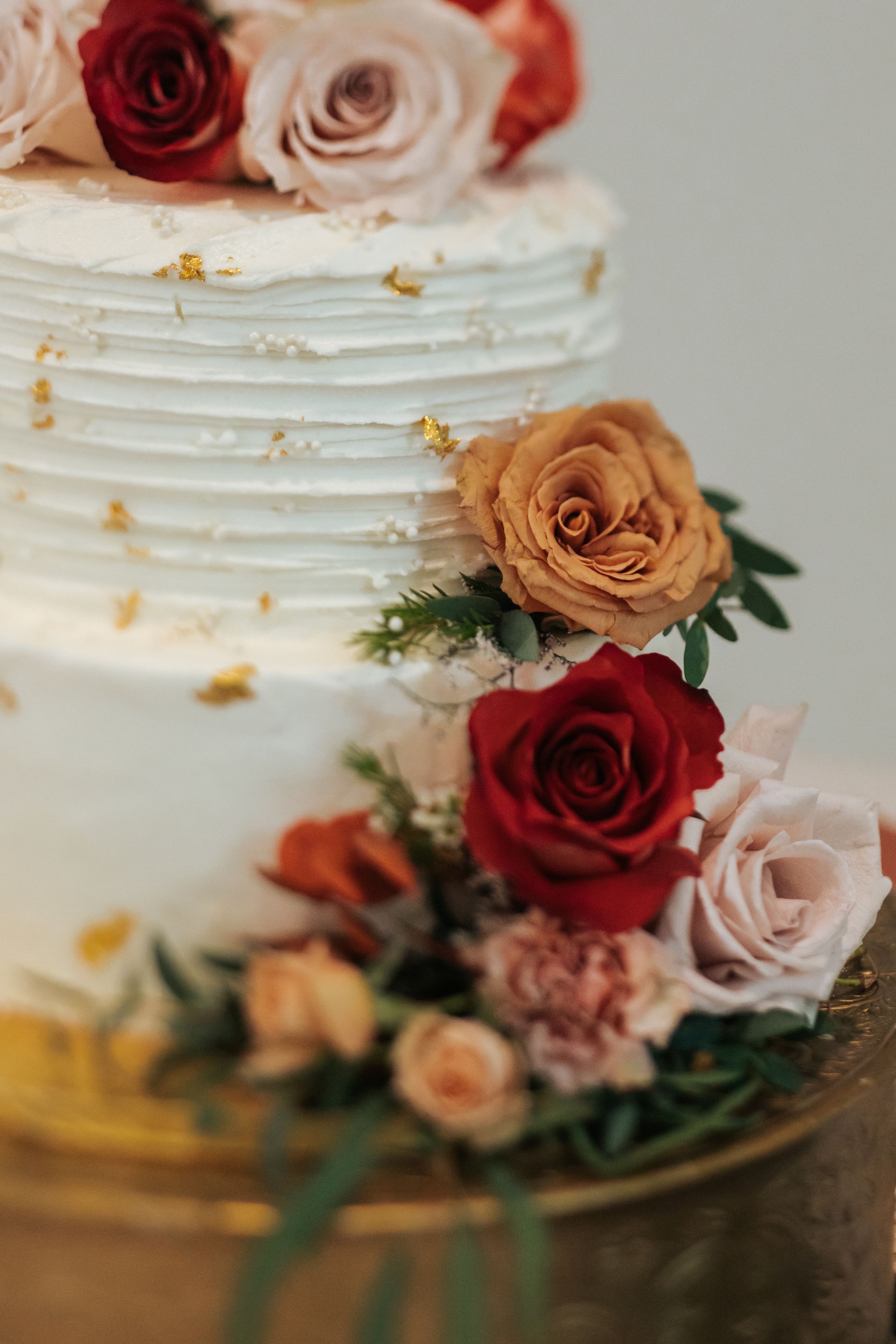 Close-up of an elegant white wedding cake decorated with red and blush roses and gold accents on a gold plate.