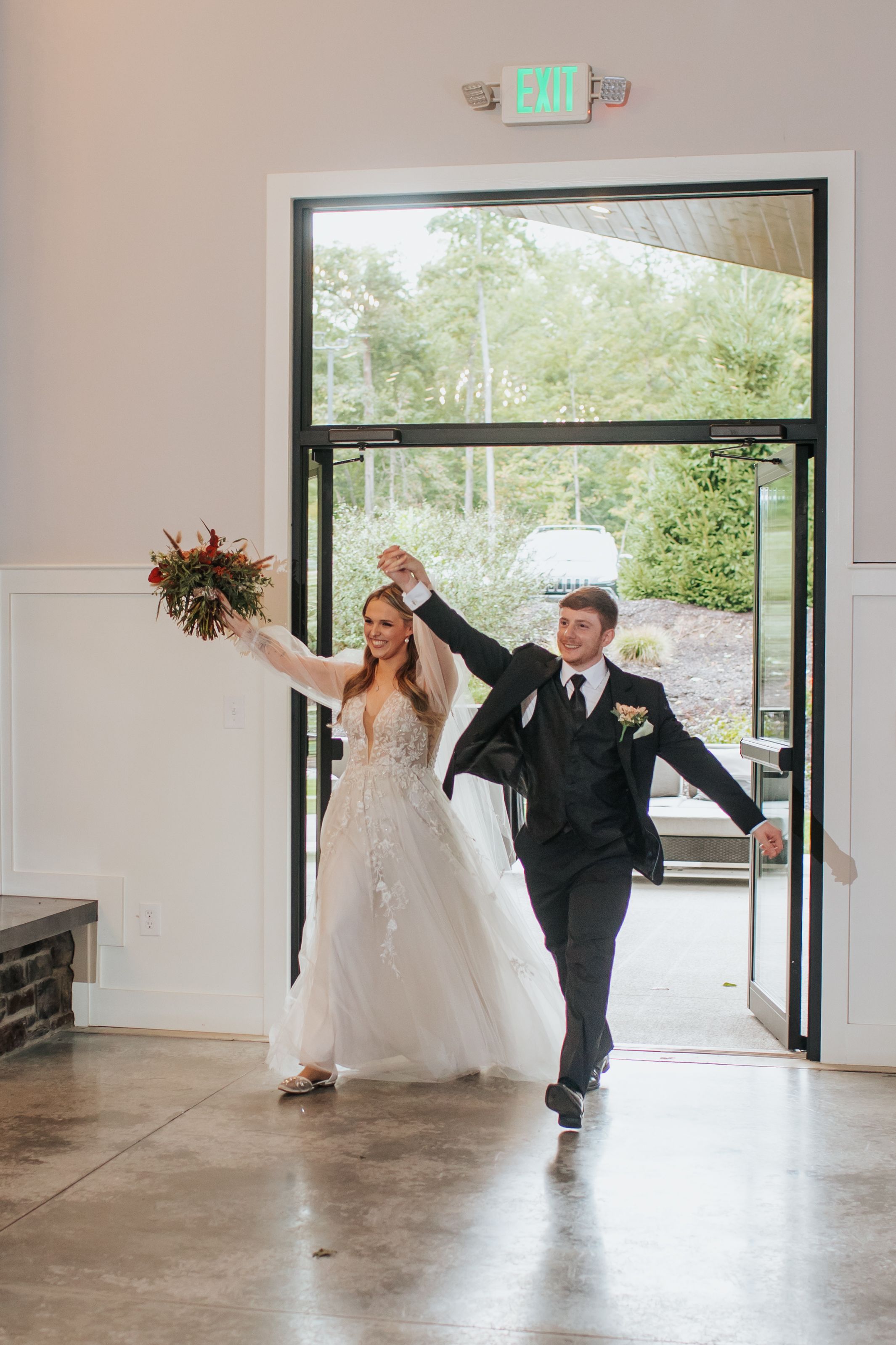A newlywed couple dances and spins joyfully through an empty indoor venue with large windows.
