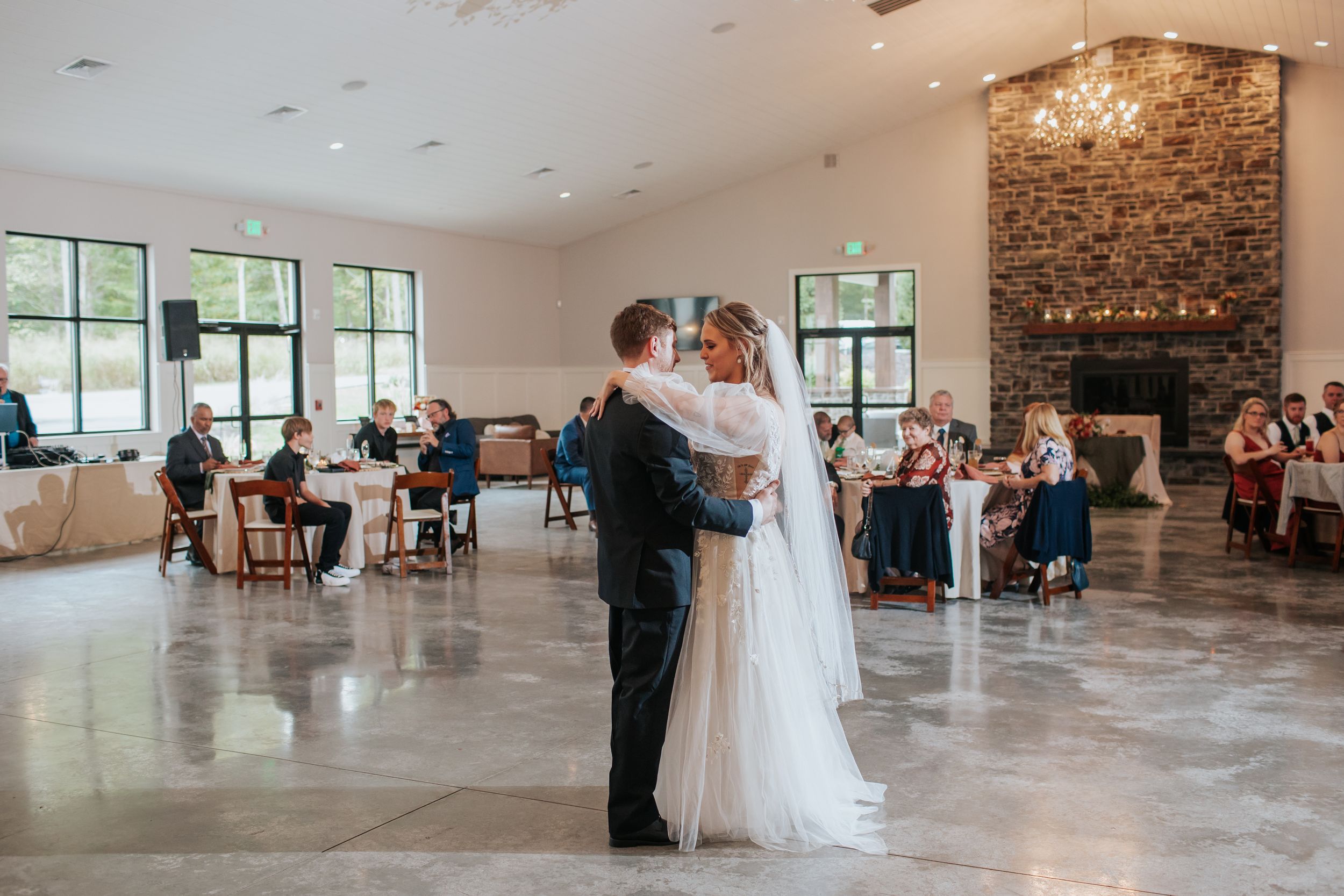 Wedding guests watch as a couple shares their first dance in an elegant industrial-style venue with brick walls.