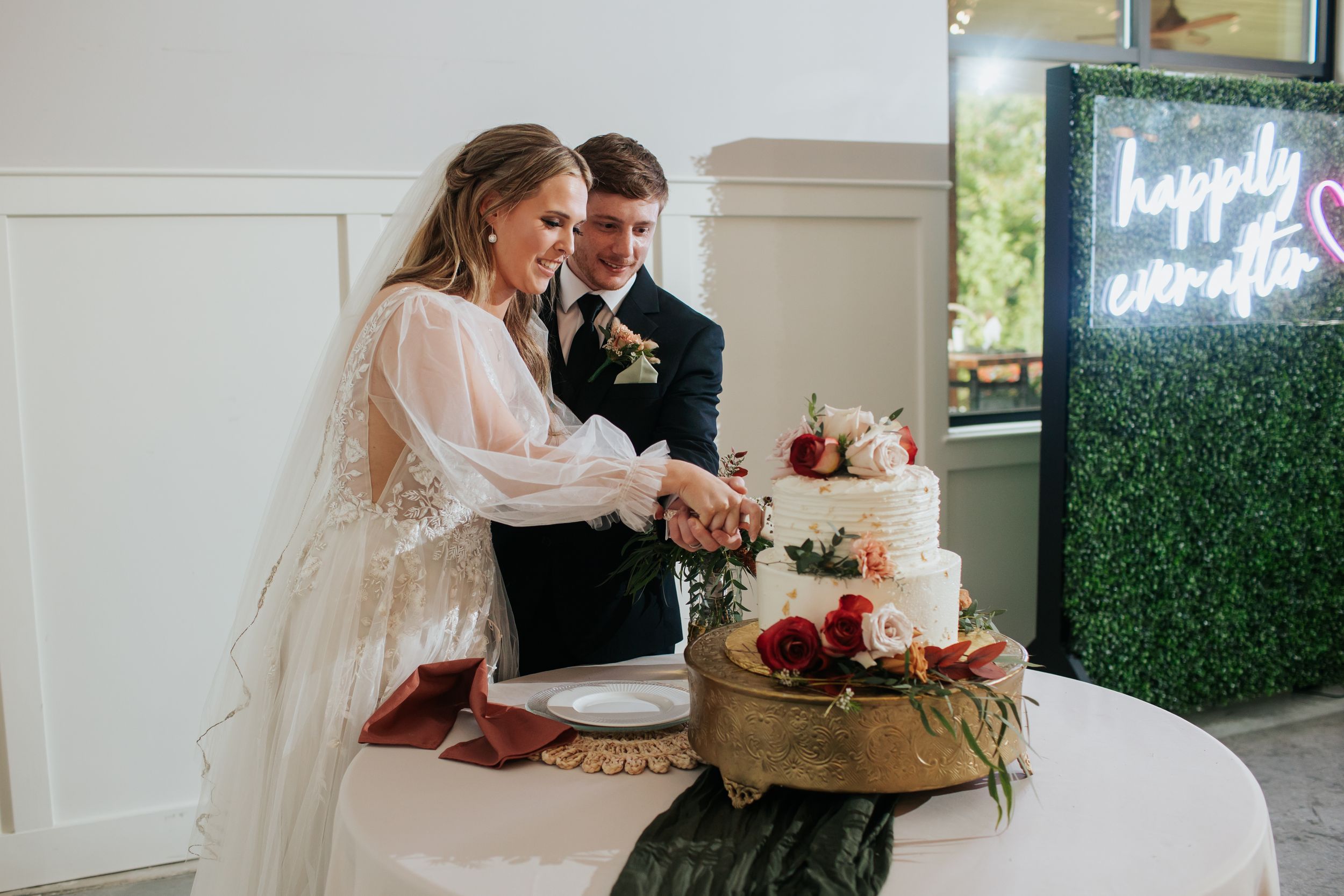 A couple in wedding attire cuts their elegant three-tiered cake together in front of a greenery wall with neon signage.