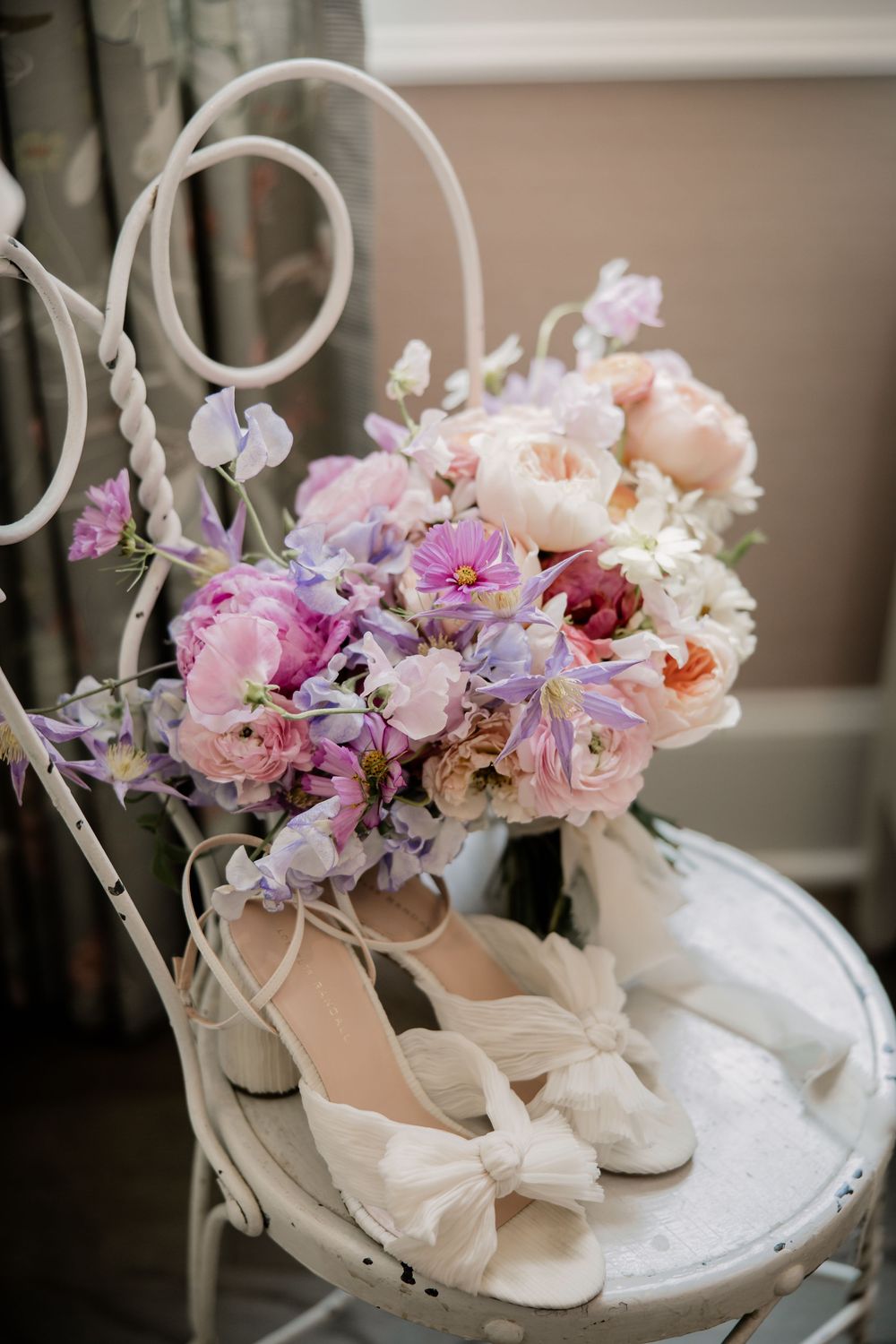 Delicate pink and purple floral arrangement with bridal shoes on vintage white chair.