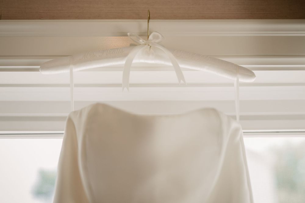 A classic white wedding dress hangs elegantly on a white hanger against a bright window.