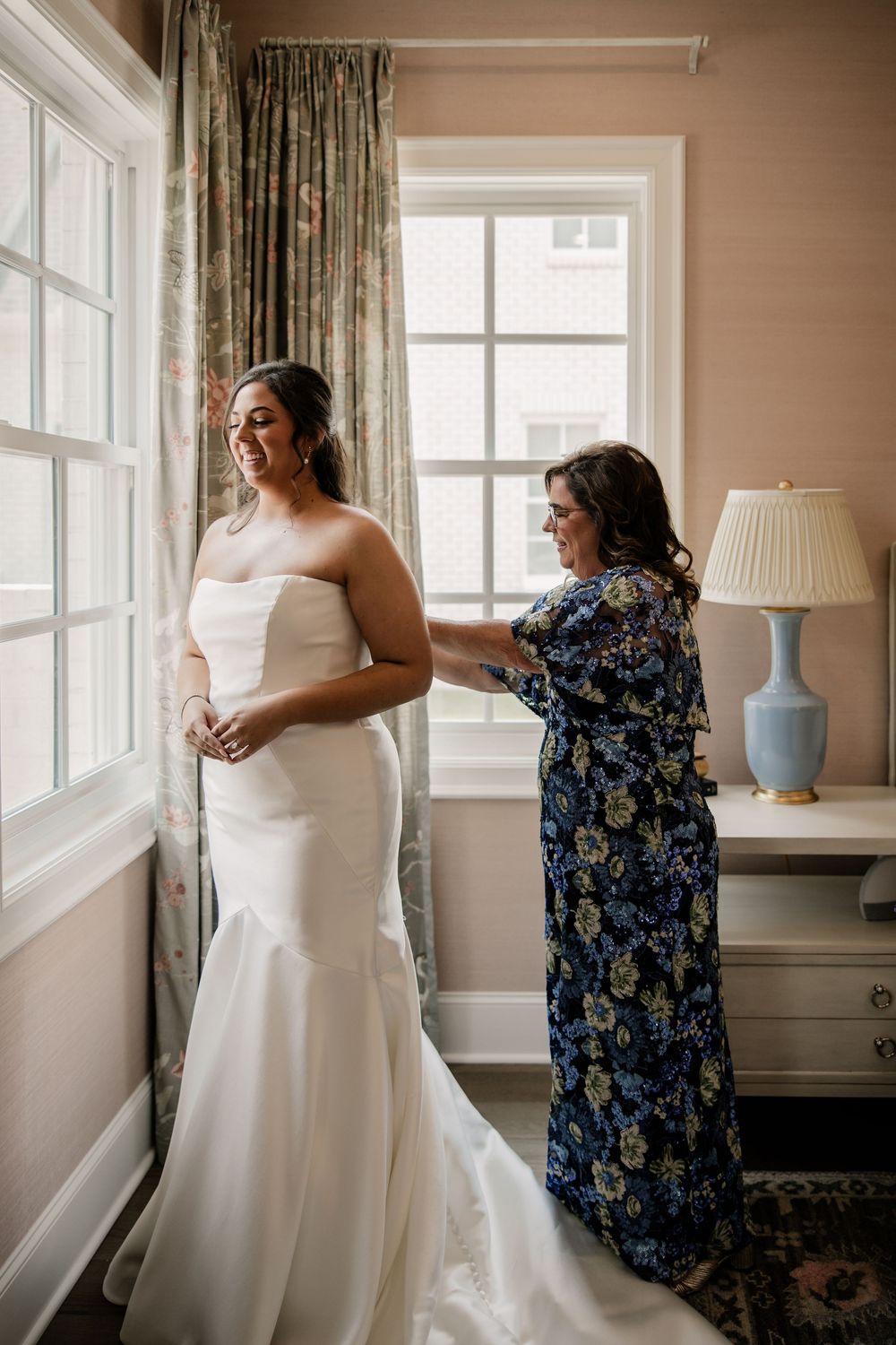 A wedding dress preparation scene in a sunlit room with floral curtains.