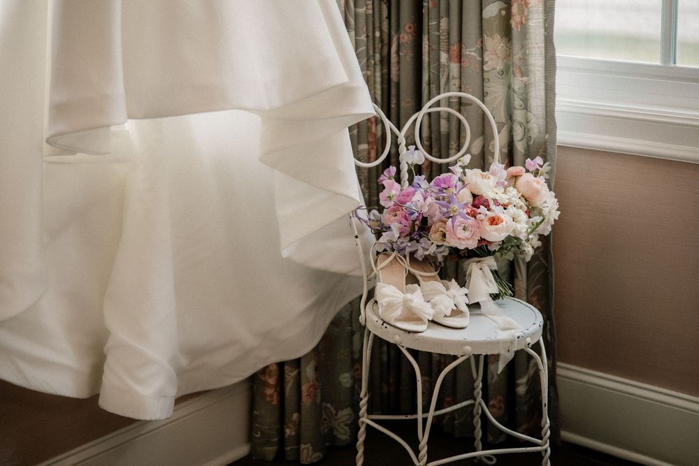 Bridal preparation room with white wedding dress hanging near window and floral arrangements.